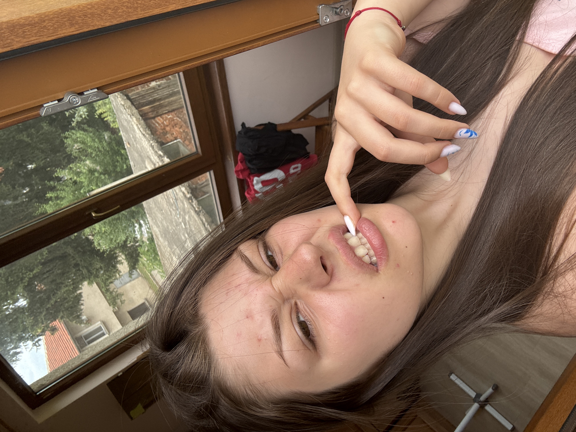 A young woman is in the foreground, her face angled towards the camera with a playful or mischievous expression. She is touching her lips with her right index finger, and her teeth are visible. Her hair is long and dark brown, falling around her face and shoulders. She is wearing a pink top, and a red string bracelet is visible on her left wrist. Her fingernails are painted white with blue abstract designs.
The scene appears to be indoors, near a window. The window frame is made of wood, and a metal latch is visible on the top of the frame. Through the window, trees and buildings can be seen, suggesting an outdoor environment. There is a reflection in the window, showing a pile of clothes, including a red shirt with white lettering and a black t-shirt. The lighting suggests it is daytime, possibly late afternoon due to the angle of the light. The overall mood is casual and lighthearted. There is no visible text in the image.