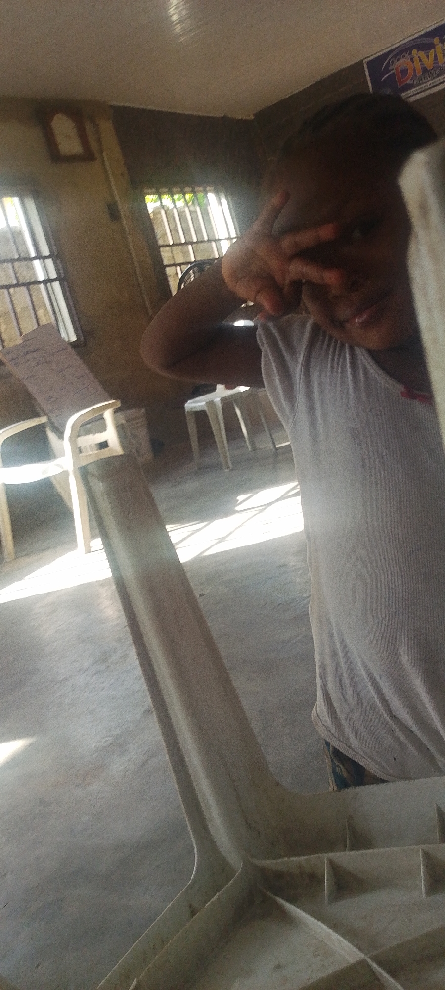 A child stands indoors, obscuring part of the view with a "V" sign over one eye. The location appears to be a room with barred windows. There are plastic chairs visible. The location is claimed to be Lokoja, Nigeria, but this is not verifiable from the image alone.