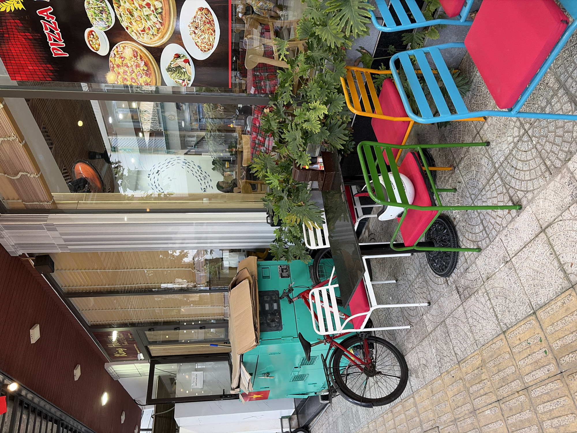The image shows an outdoor seating area in front of a restaurant in Đà Nẵng, Vietnam. A bright turquoise generator is parked on the sidewalk, covered with cardboard. A red bicycle is leaning against the generator. Several colorful metal chairs with red cushions surround a black outdoor dining table. Plants are arranged around the seating area. In the background, through the restaurant window, people can be seen dining. A poster advertising pizza is visible on the window. The sidewalk is paved with textured tiles, and a ramp with tactile paving is present.