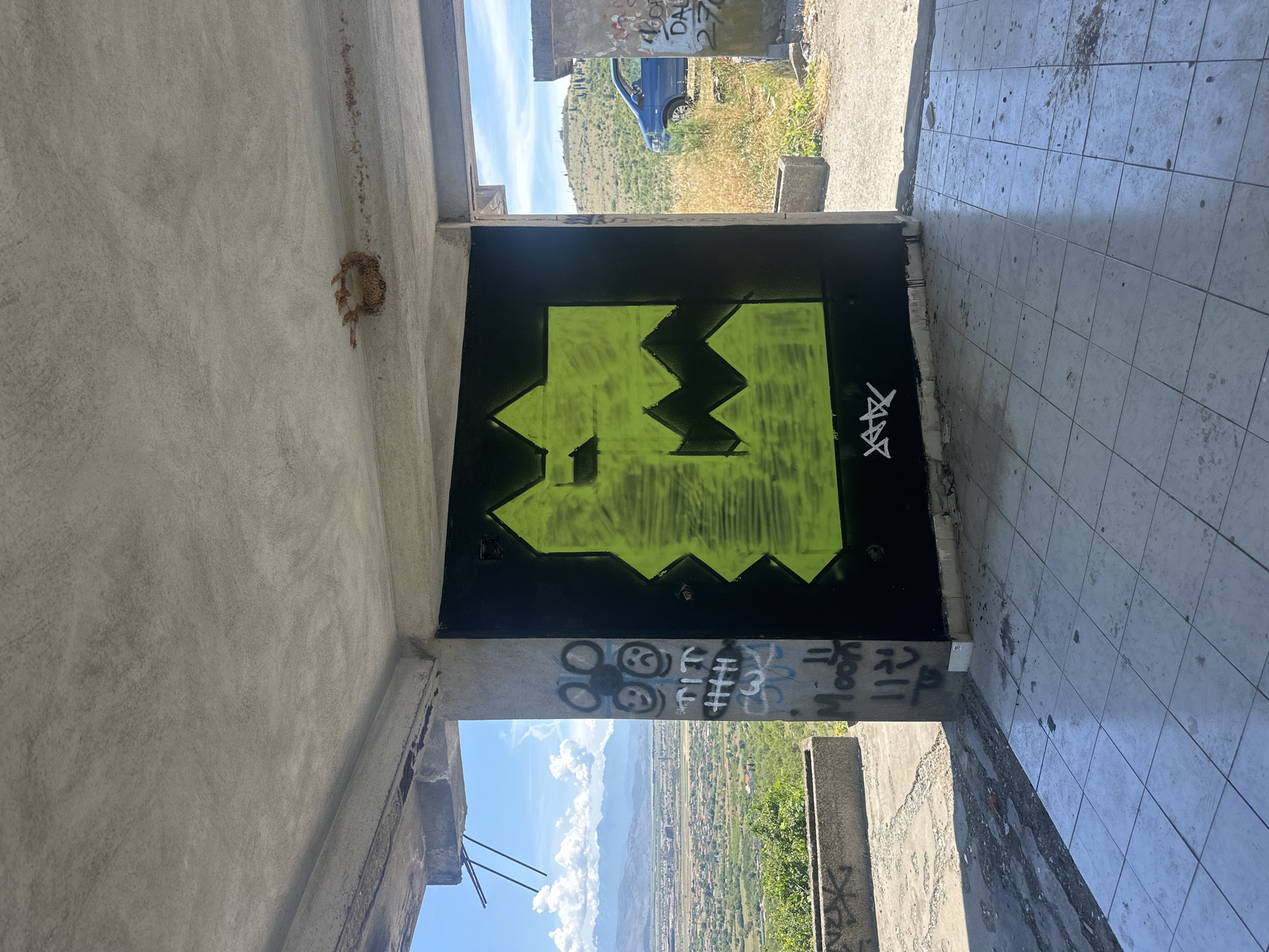 This image depicts the interior of a partially constructed or abandoned building in Gornji Kokoti, Montenegro. The foreground is dominated by a tiled floor with some debris. On the back wall, a large, jagged graffiti tag in bright lime green spray paint is prominently displayed against a black background. To the left of the graffiti, a concrete pillar is covered in various smaller tags and drawings, including smiley faces and what appears to be the word "Moon".
The building appears to be open to the elements, with views of a rural landscape in the background. Rolling hills covered in vegetation, a town or village, and mountains under a bright blue sky with scattered clouds are visible. A blue car is parked outside the structure to the right. The overall scene suggests a state of disrepair and neglect, common in abandoned or unfinished structures.