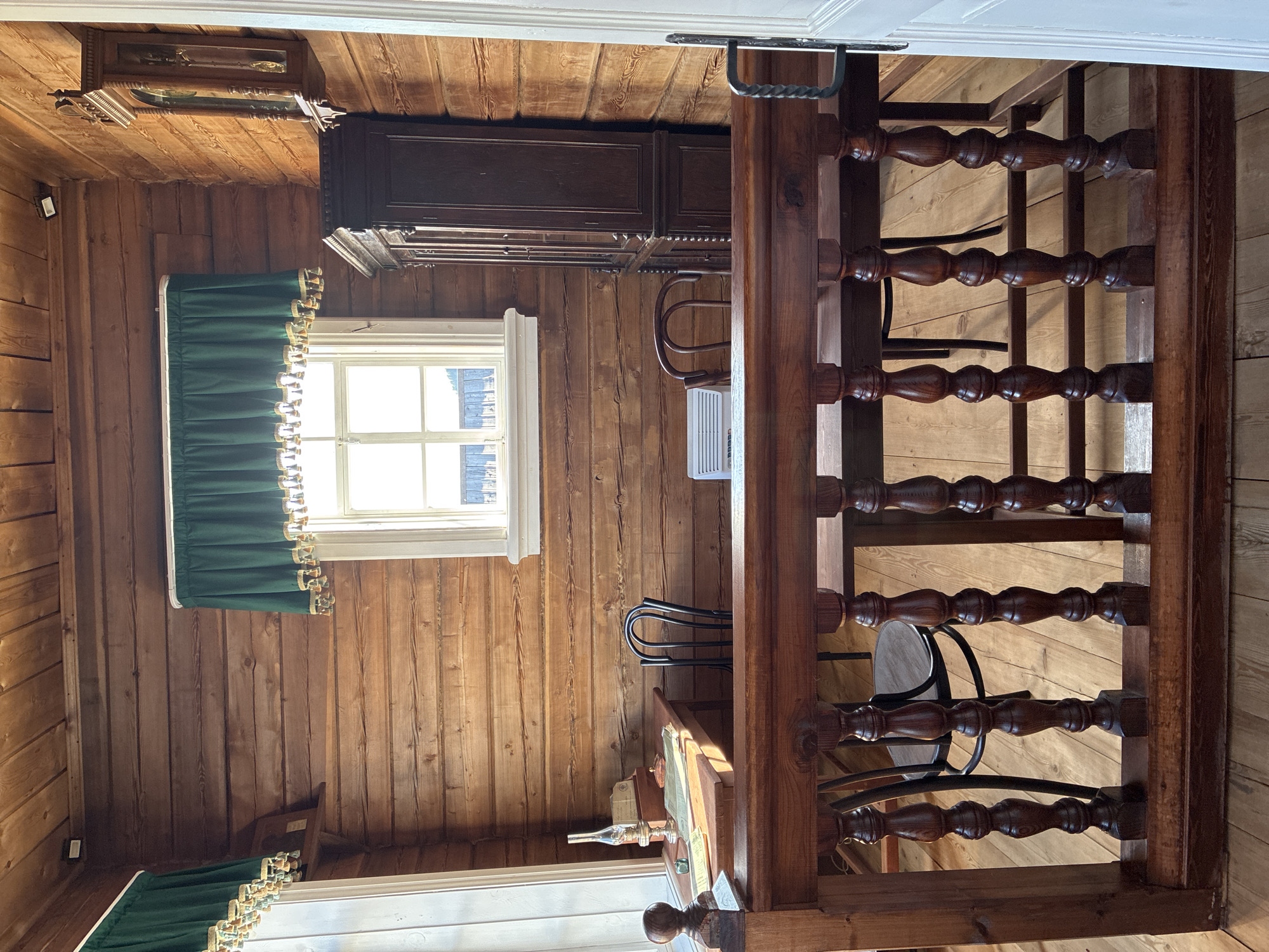 The image depicts the interior of a room in a wooden log building, likely a museum exhibit at the Museum of Wooden Architecture in Taltsy, Russia. The walls and ceiling are constructed of horizontal wooden logs.
In the foreground, a wooden railing with turned balusters separates the viewer's perspective from the main area of the room. The floor is also made of wooden planks.
Behind the railing, a wooden desk is visible on the left side of the room, near a window. On the desk are several papers, a glass bottle, and what appears to be an oil lamp. A vintage-style chair with a curved back is positioned behind the desk.
To the right of the desk, a large, dark wood cabinet with glass doors stands against the wall. Above the cabinet, a pendulum clock with an ornate wooden casing is mounted on the wall. A second chair, also with a curved back and made of metal, is visible near the cabinet. A white, rectangular air conditioning unit is placed on a table in front of the cabinet.
A window with a white frame and multiple panes is centrally located on the back wall. A green curtain with decorative tassels hangs above the window. The window looks out onto an outdoor scene, possibly a fence and trees. Another window, partially obscured by the foreground railing, is on the left wall.
The overall impression is that of a preserved historical interior, likely a study or office from a past era. The lighting in the room is a mix of natural light from the windows and interior light from unseen sources, casting soft shadows.