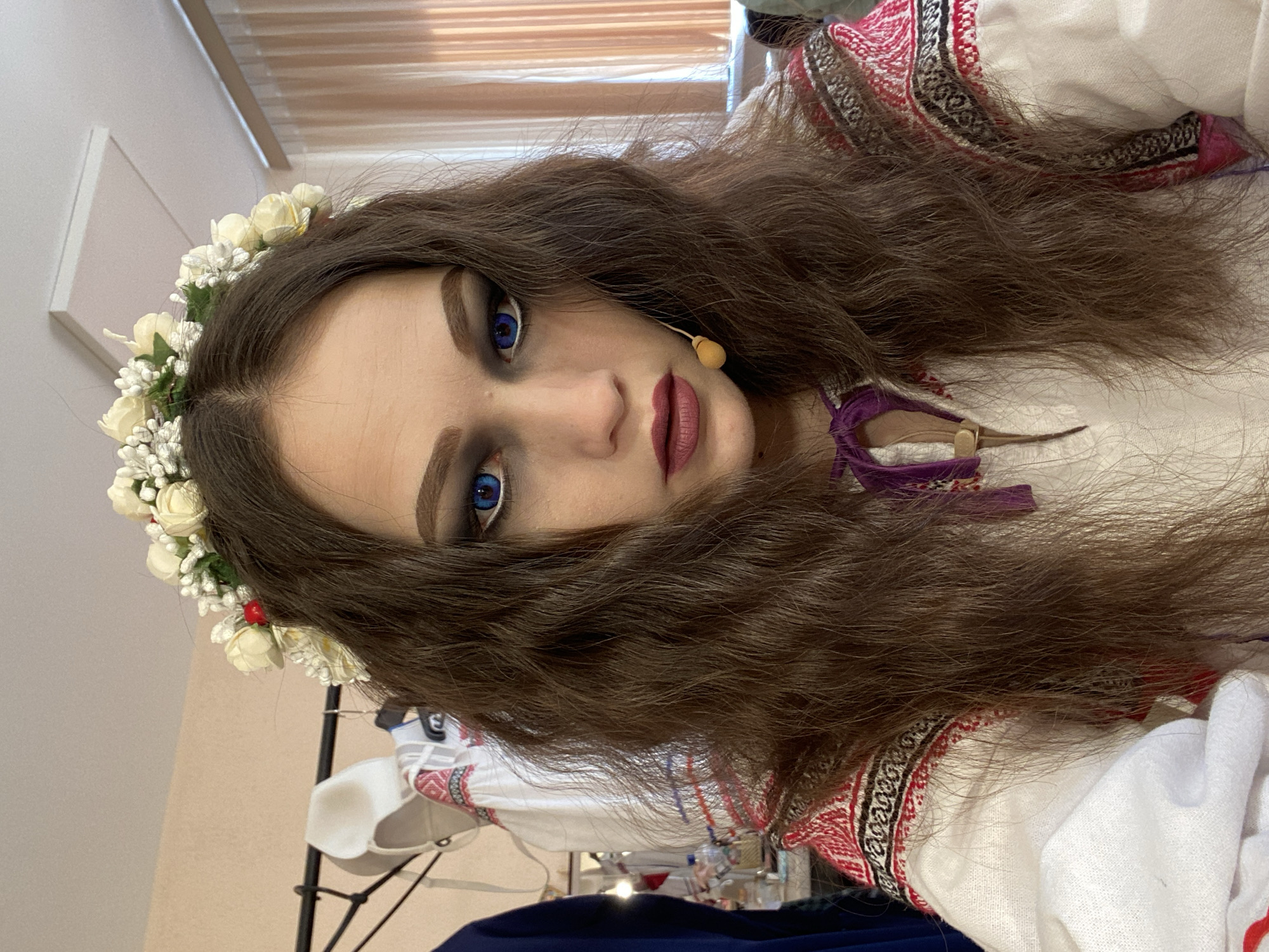 The image shows a woman with long, wavy brown hair adorned with a floral crown. She has fair skin, dramatic makeup including dark eyeshadow and plum lipstick, and strikingly blue eyes. She is wearing a white traditional shirt with embroidered patterns in red, black, and white around the collar and sleeves. A purple tie is visible at the collar.
The background is slightly out of focus but suggests an indoor setting, possibly a dressing room or studio, with beige walls and partially visible furniture and equipment. The location in the city of Chaikovsky, Russia, is not directly identifiable from the visual information in the image.