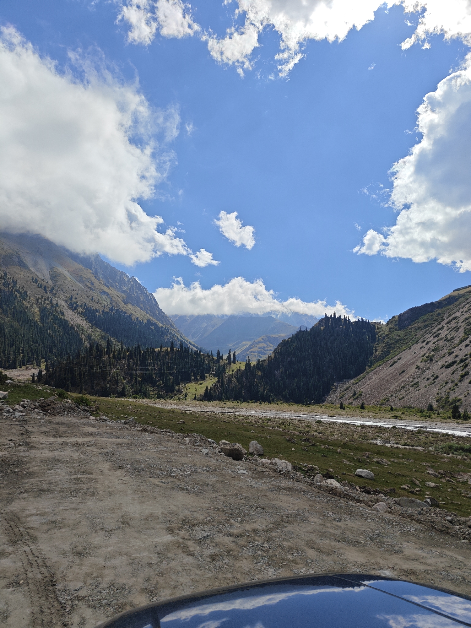 The image depicts a panoramic view of a mountain valley in Kyrgyzstan. The foreground is dominated by a dirt road with tire tracks, suggesting it's a passage for vehicles. To the right, the hood of a dark blue vehicle is visible, reflecting the sky and clouds. The middle ground features a lush green meadow dotted with large rocks, leading to a winding river. Further back, a dense forest of coniferous trees covers the slopes of the mountains. The mountains themselves are rugged and steep, with some peaks partially obscured by dramatic white clouds against a bright blue sky. The overall scene conveys a sense of natural beauty and remoteness.
