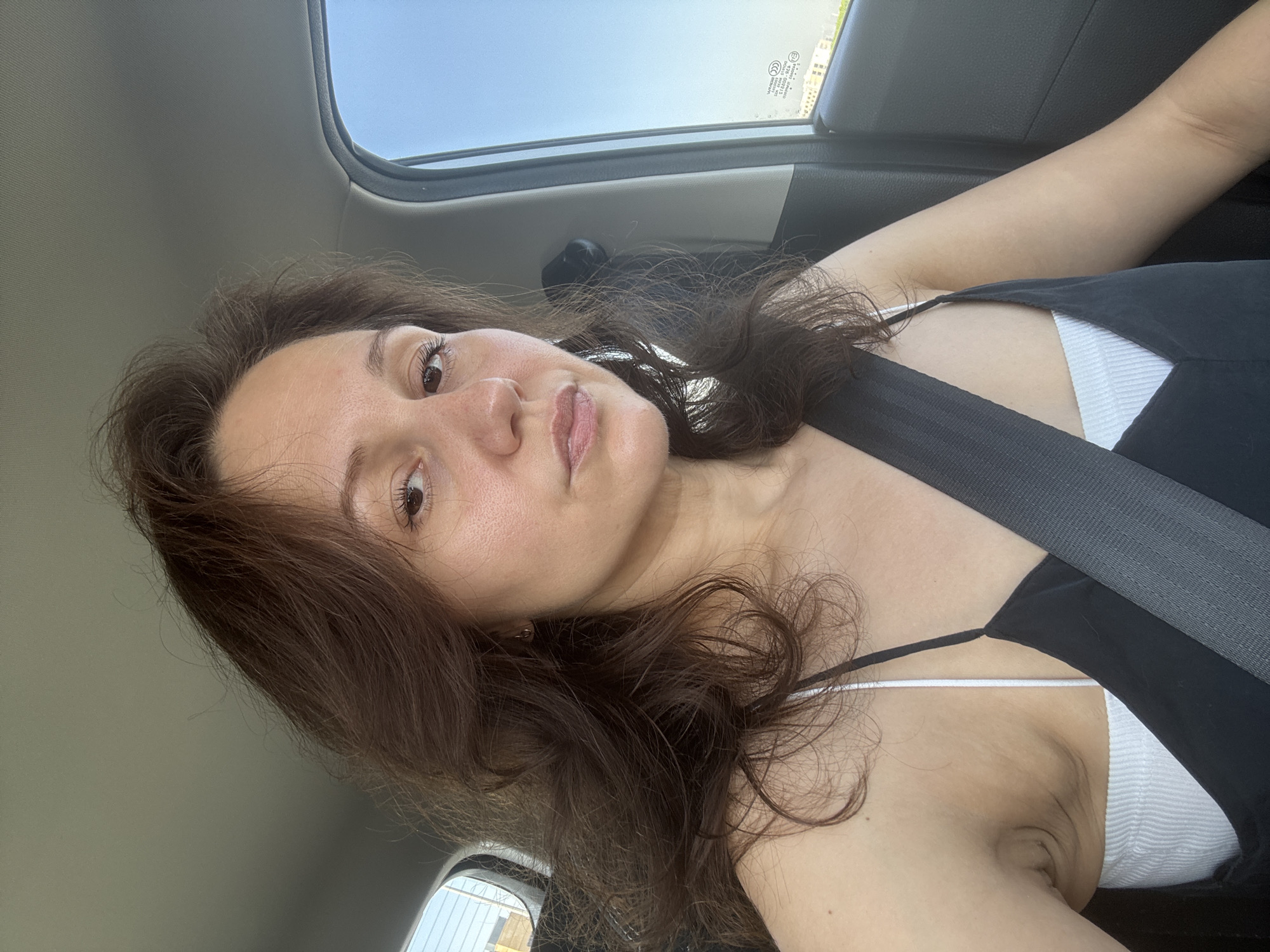 A woman with long brown hair sits in a car wearing a black top with white straps and a seatbelt. The scene takes place inside a vehicle, and the location cannot be determined from the image.
