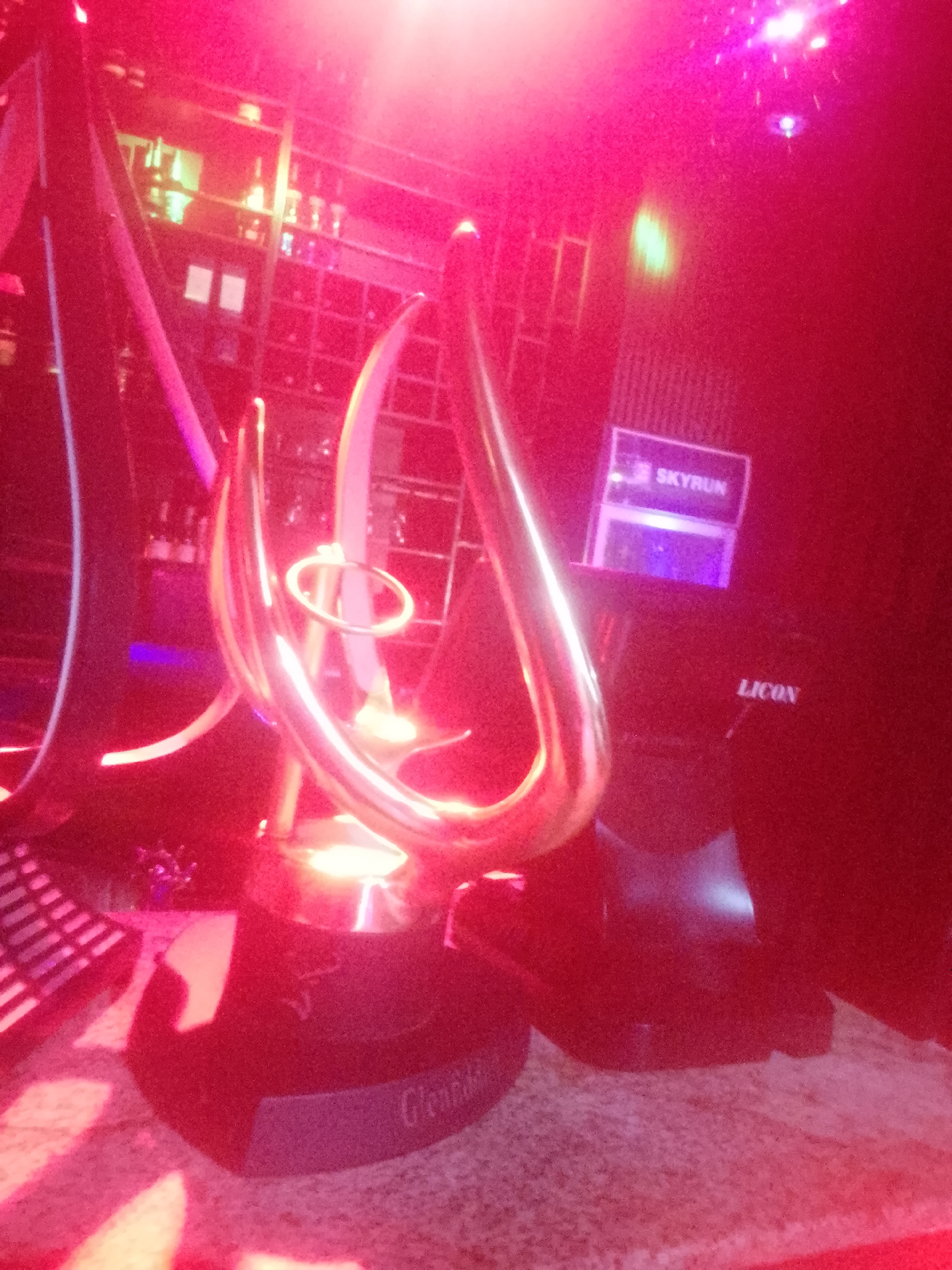 The image depicts a dimly lit indoor scene, likely a bar or club, bathed in vibrant red and pink lighting. The focus is on a trophy-like object with a golden, curved structure, resting on a dark pedestal. The pedestal has text that appears to read "Glenfiddich" on its base. Behind the trophy, a black object with a sign that says "SKYRUN" is visible, with another sign below it reading "LICON". The background shows shelves filled with bottles, suggesting a bar setting, and a large dark structure that could be a stage or a wall. The overall ambiance is one of an evening event or celebration. The city of Howayi, Nigeria is not discernible from the image itself.