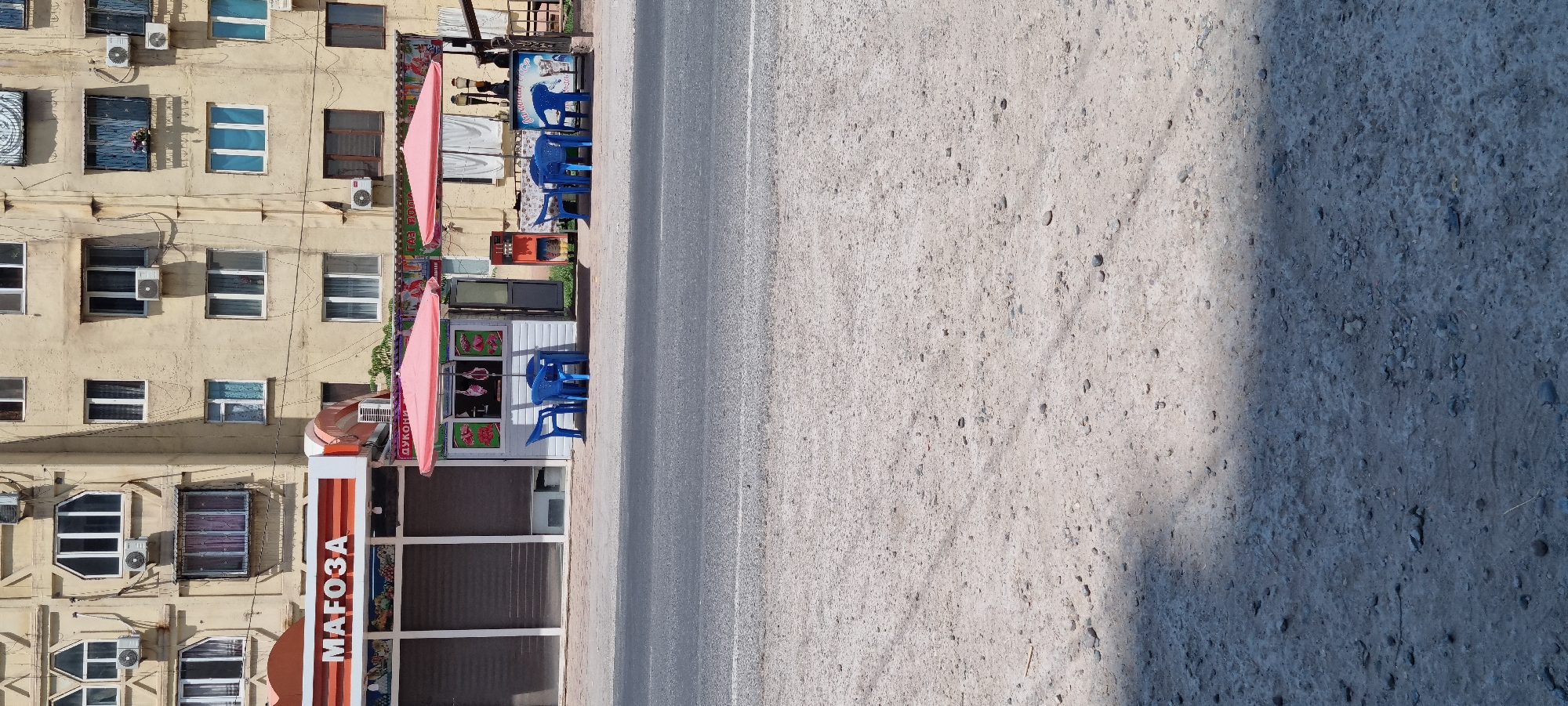 The image shows a street scene in the city of Ismoili Somoni, Tajikistan. In the foreground, there is a paved road with a gravel shoulder. In the mid-ground, there are two small food stalls with pink awnings. The stall on the left appears to sell meat, with carcasses hanging on display. The stall on the right has tables and chairs set up outside, with signs that appear to advertise drinks and possibly ice cream. In the background, there is a multi-story building with a yellowish facade, featuring numerous windows and air conditioning units. The building's windows are a mix of styles, some with visible curtains. The overall scene depicts a common urban commercial area, likely a busy street with small businesses catering to passersby.