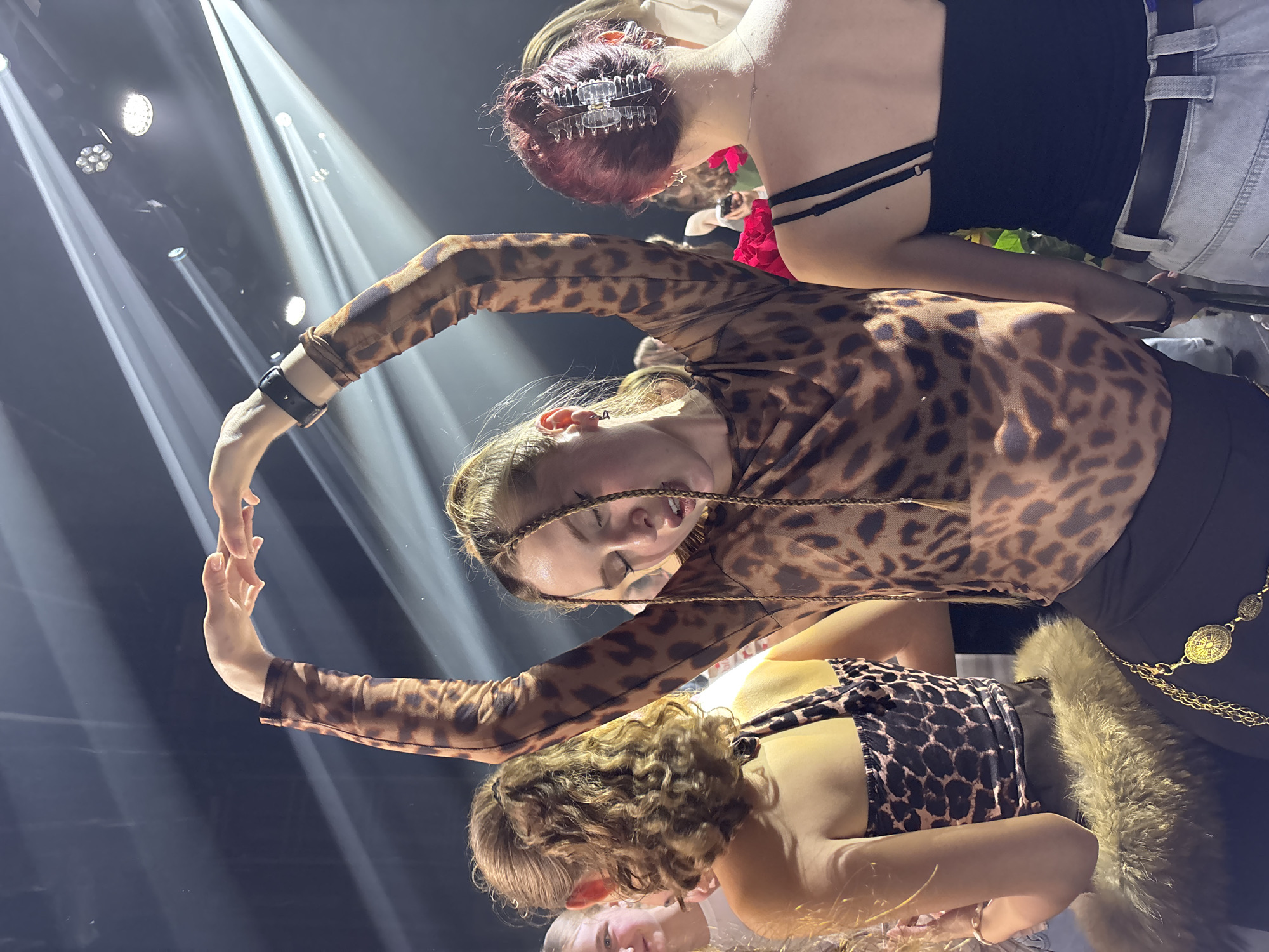The image shows three women on a stage under bright stage lights, likely in a performance setting. The woman in the middle wears a leopard print top and is positioned so that her arms create a semi-circular shape. Below her is a blonde woman in a similar leopard print top, with a fur-like accessory. Above the central woman is a woman with reddish-brown hair, wearing a black top, light jeans and a belt. The location may be in Perm, Russia.