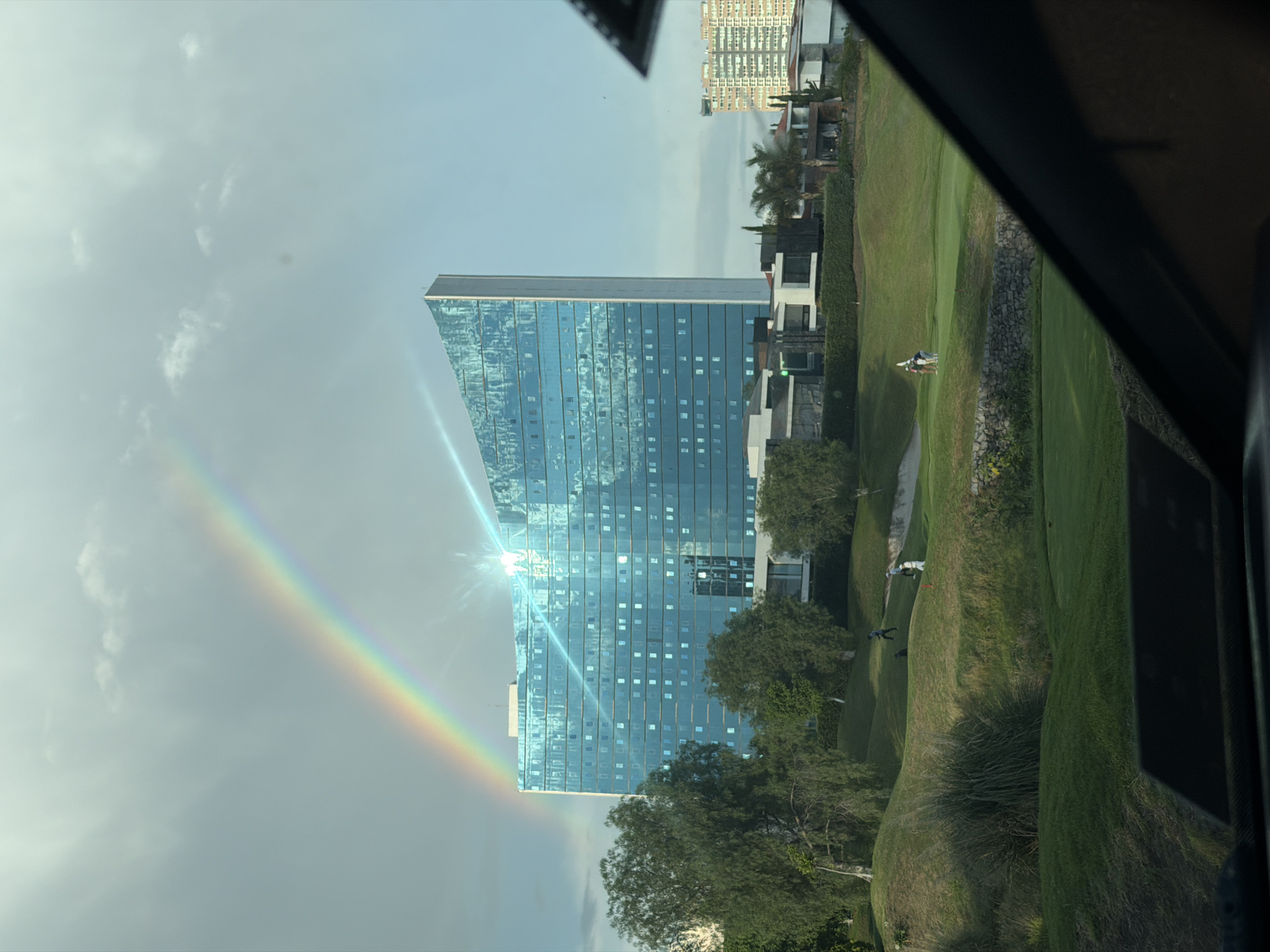 The image displays an outdoor scene in San Bernardino Tlaxcalancingo, Mexico, viewed from within a vehicle, evidenced by the dark dashboard and window frame partially obscuring the lower left and right corners. The central feature is a large, modern, multi-story building constructed with reflective blue glass, exhibiting a curvilinear or angular architectural design that culminates in a sharp point on its right side. A significant sun flare or bright reflection emanates from the upper right section of this building. Arcing across the cloudy sky, a vivid rainbow is visible, with its base appearing behind the left side of the building and extending over its top towards the right.
In the midground, a meticulously maintained green golf course with undulating terrain spans the width of the frame. Several trees with light-colored trunks and green foliage are scattered across the course. Multiple individuals are present on the golf course: a group of three is visible on a rise on the left, one figure in dark attire and two in lighter clothing, appearing to be golfers or caddies. Further to the right, two additional figures are standing near a red flagstick, one in a light shirt and dark pants, and another in white attire. A small, dark, indistinct figure is also visible on a greenspace between these groups. A low stone wall forms a boundary across a section of the golf course.
Behind the golf course, a cluster of contemporary residential structures with flat roofs, featuring contrasting white and dark facades, is visible. One residence on the left exhibits a distinct green light or architectural feature. Palm trees are also interspersed among these residences. In the far background, on the right side of the image, another taller, multi-story residential building with a more uniform, grid-like facade is discernible under the partially cloudy, light blue sky.