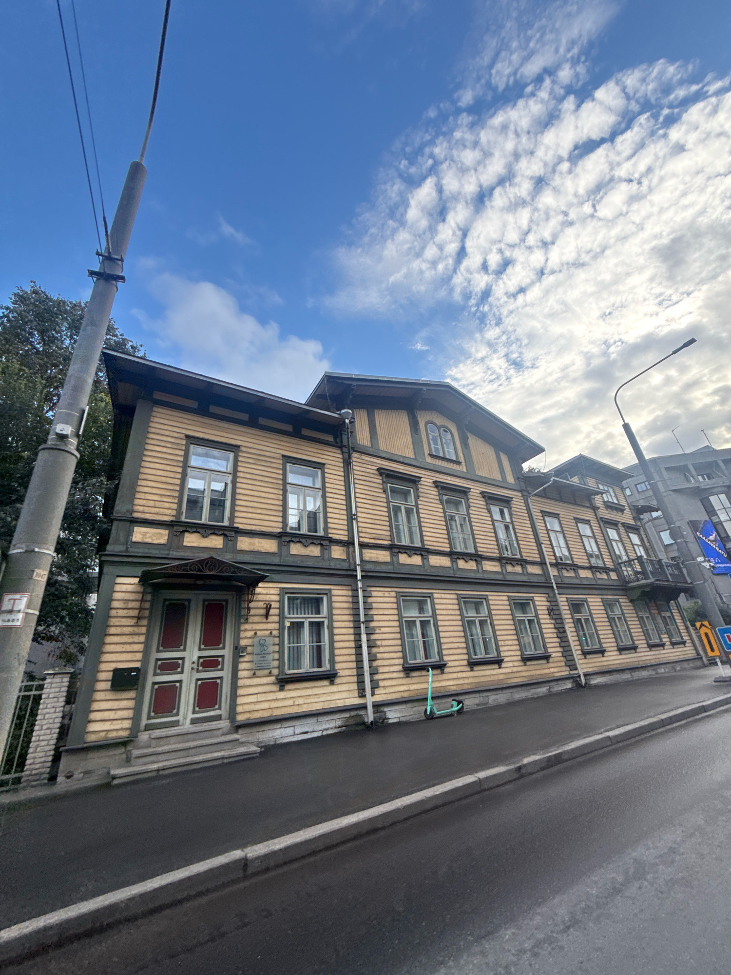 The image depicts a street scene in Tallinn, Estonia, focusing on a narrow, wooden, two-story building with a light yellow facade and dark green trim. The building features numerous windows with dark frames and what appear to be red doors. A covered entrance with decorative metalwork is visible at the ground level. To the left of the building, a tall, grey utility pole with wires extends upwards into a blue sky dotted with white, wispy clouds. In front of the building, a wet asphalt sidewalk runs alongside a street. A light green electric scooter is parked on the sidewalk near the building. Further down the street, modern buildings can be seen, including a taller grey structure on the right. A street lamp is positioned on the right side of the image. The overall impression is a blend of historical architecture and modern urban elements under an overcast sky.