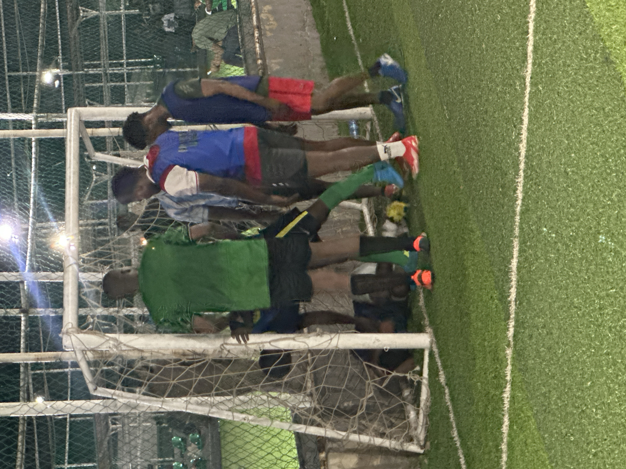The image depicts an outdoor artificial turf soccer field at night, illuminated by artificial lighting. A group of young men, presumably soccer players, are gathered around a soccer goal on the left side of the frame. One individual is on the ground inside the goal net, while others stand around him. The players are wearing various athletic attire, including shorts, jerseys, and soccer cleats in shades of green, blue, red, and white. The background shows a chain-link fence and further beyond, hints of urban structures and possibly other spectators, suggesting a public or semi-public sporting venue within Abuja, Nigeria. The overall atmosphere suggests a moment of pause or concern during a soccer game.