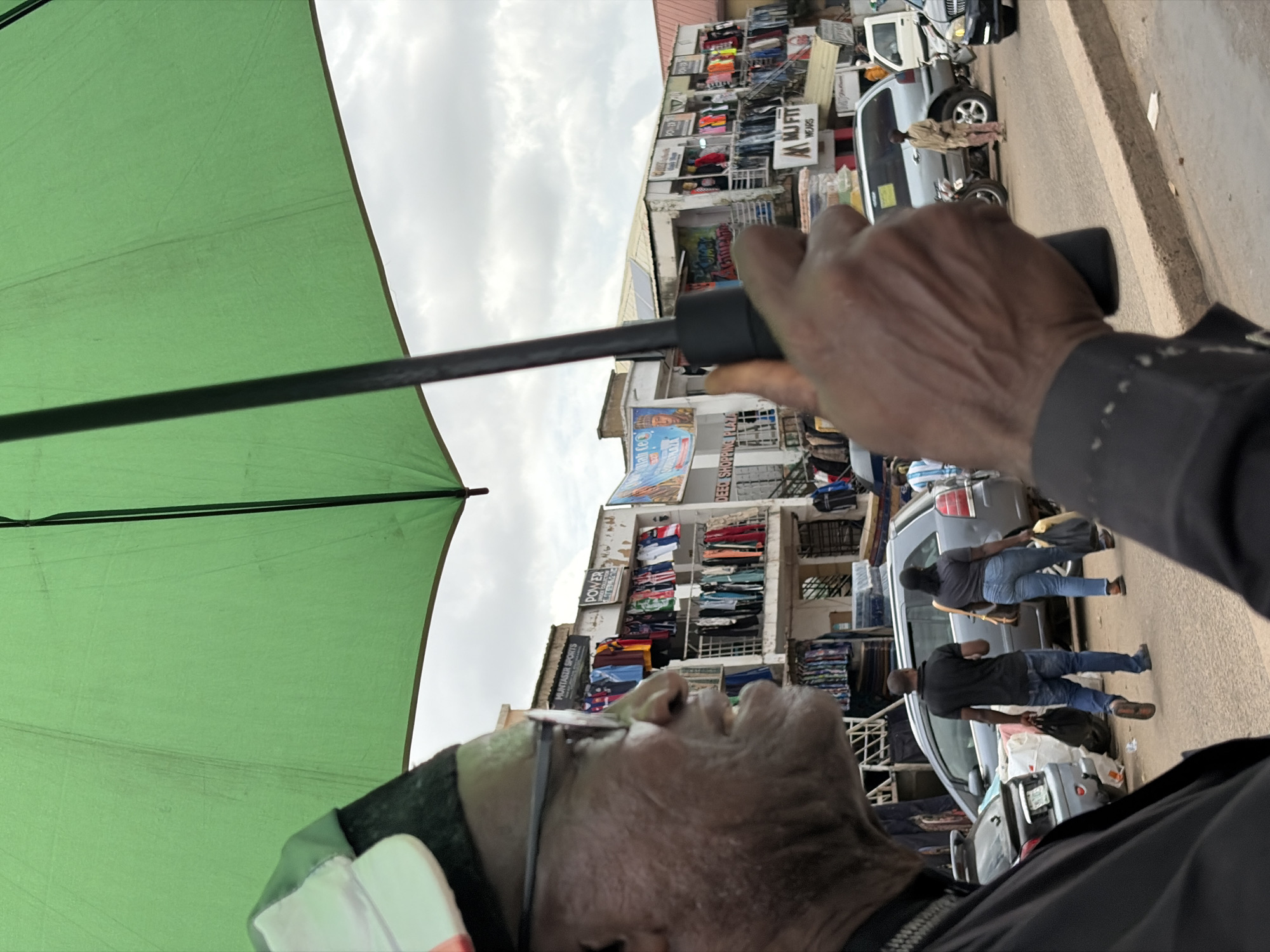 In an outdoor market scene in Jos, Nigeria, a person, largely obscured by a large green umbrella, is holding the umbrella's handle in their right hand. The person wears glasses and a cap. In the background, a row of shops displays clothing items. A silver SUV, a black car, and a white van are parked on the street. Two individuals are walking away from the camera carrying shopping bags. The sky is overcast.