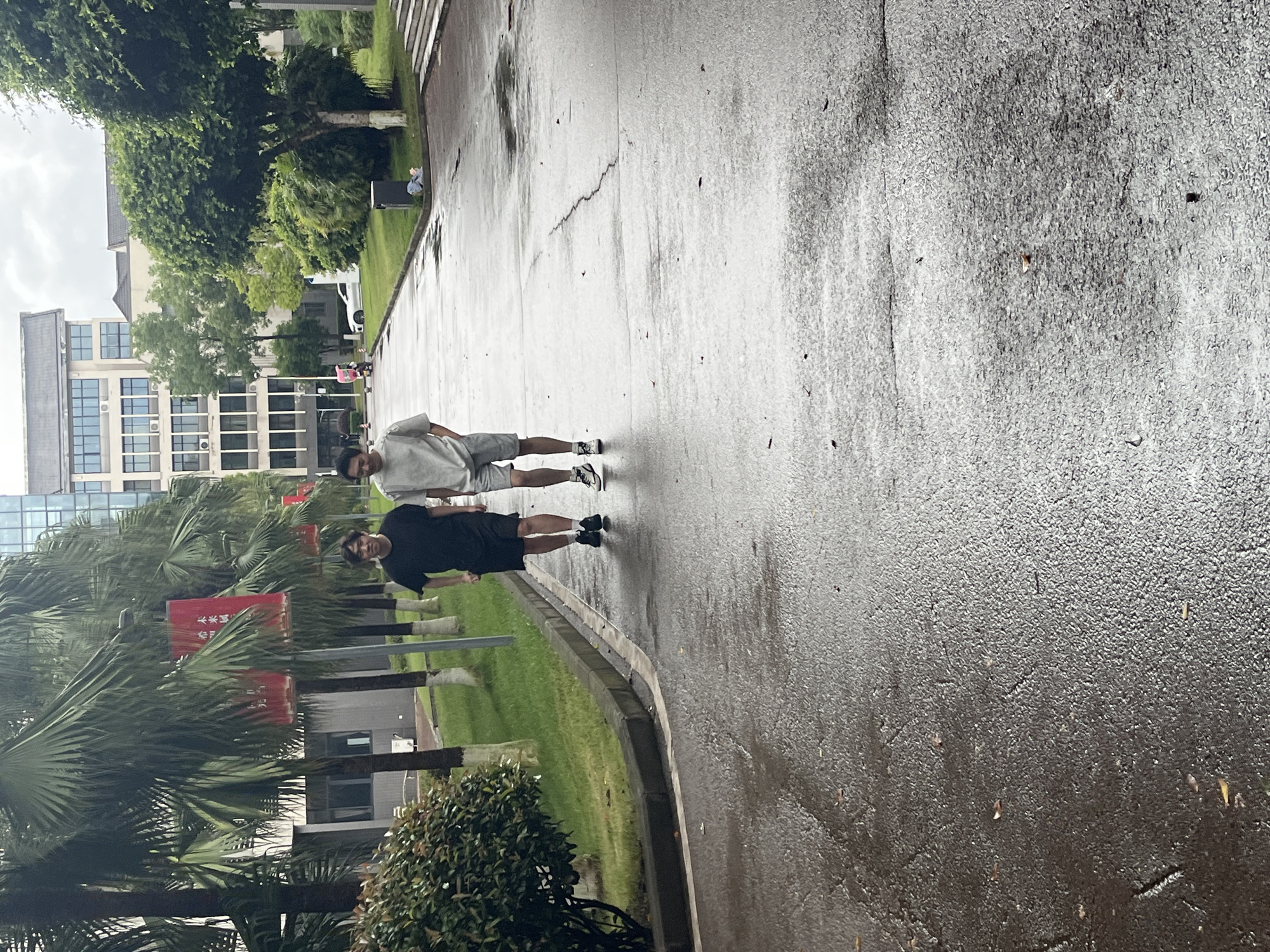 Two young men are walking on a wet asphalt path lined with palm trees and grass. The man on the left wears a black t-shirt and black shorts, while the man on the right wears a light-colored t-shirt and gray shorts. Both are wearing athletic shoes. In the background, there are multi-story buildings, some with visible windows and air conditioning units. A white car is parked in the distance. The environment appears to be a campus or park area, given the landscaping and buildings. The presence of wet surfaces suggests recent rain. This scene is likely located in the city of Jiangjin, China, given the context.