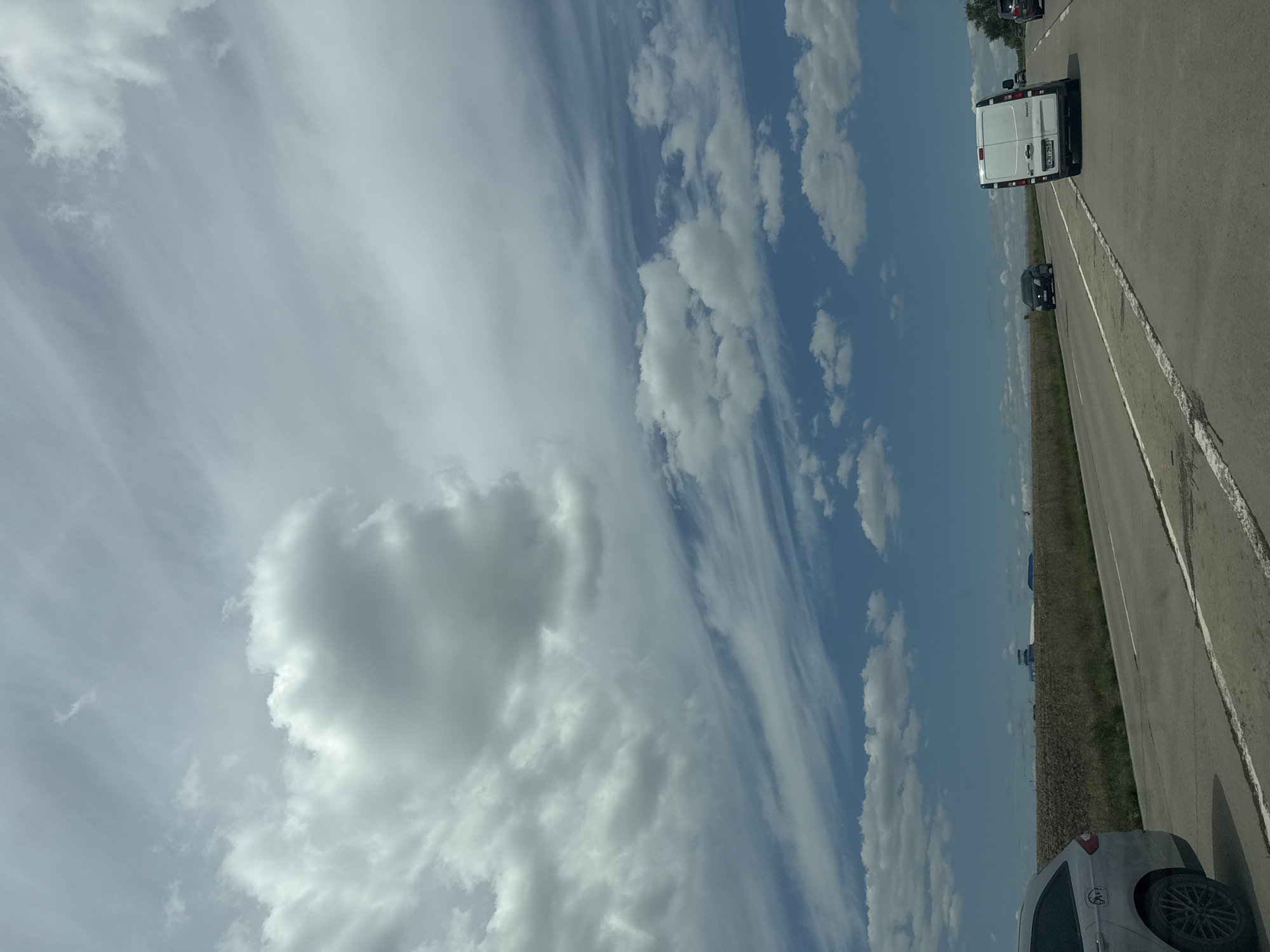 The image depicts a highway scene in Horesti, Moldova, under a cloudy sky with patches of blue. In the foreground, a silver SUV is parked on the left side of the road. Further down the road, a white van with a license plate reads "VWJ 263" is driving in the right lane. Other vehicles are visible in the distance. The landscape features a dry, grassy field bordering the highway, with a few trees on the right side. In the far distance, a building with a tall structure, possibly a tower, is visible against the horizon. The overall impression is one of a typical roadside environment on a partially overcast day.