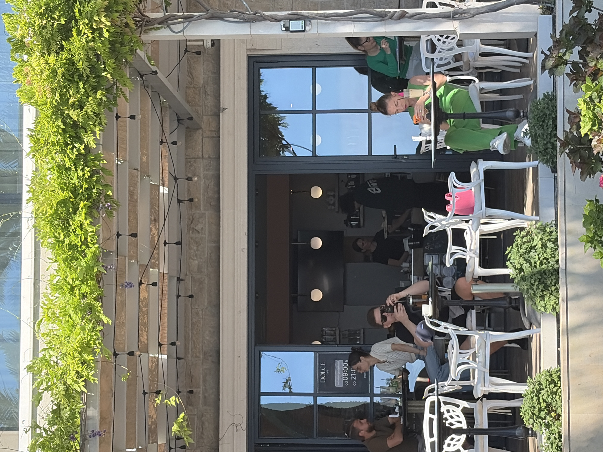 The scene is set at an outdoor cafe located in Bečići, Montenegro, as evidenced by the "DOLCE CAFE" sign. The cafe has a stone wall and an awning covered with climbing vegetation and string lights. Patrons are seated at white tables with white chairs. Several people are visible: a woman in a green outfit sitting with another person, a person taking a photo, and a group of people seated at other tables. The cafe has glass doors and windows reflecting the outdoor environment. The light suggests it is daytime.