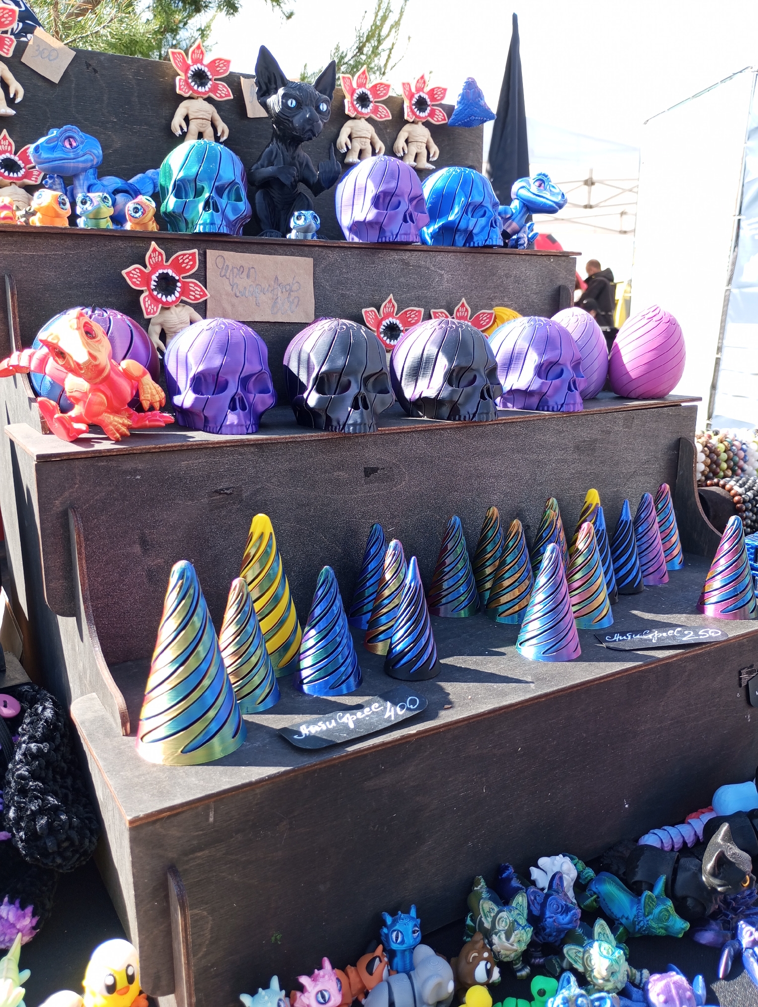 The image depicts a multi-tiered wooden display stand showcasing an assortment of handmade and 3D-printed figurines at an outdoor market. The top tier features a variety of items, including skulls in different colors (blue, purple, and black), Demogorgon figures (humanoid bodies with a flower-like head), and a stylized black cat. The middle tier primarily contains more skulls, Demogorgon figures, and a bright orange turtle figurine. The bottom tier is filled with cone-shaped objects featuring colorful striped patterns and small animal figurines. Price tags in Russian rubles are visible, with one reading "Кольца спираль 250" (Spiral rings 250) and another stating "Глазки сердец 400" (Eyes hearts 400). The location is identified as Izhevsk, Russia. The lighting suggests a sunny day. In the background, there are blurry figures and a white tent. The scene suggests a local craft market or fair.