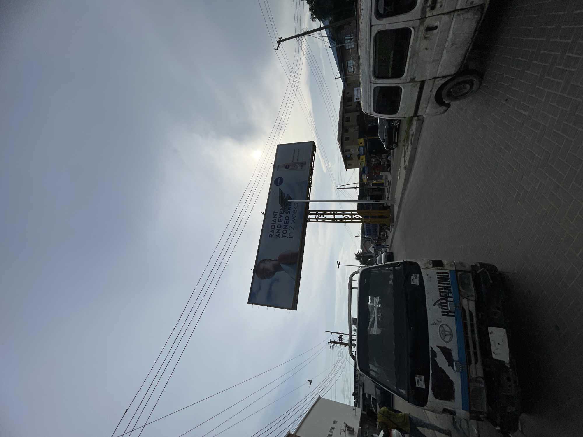 The image captures an outdoor street scene under a bright, overcast sky, likely in Ado, Nigeria. The perspective is slightly angled, looking upwards towards a large billboard and the sky, with various elements of urban infrastructure present.
In the foreground and mid-ground, two weathered white vans or minibuses are visible. The closer vehicle, on the left side, displays "HOPEFUNI" and a Toyota logo on its front-left side. A person wearing a yellow top is partially visible near the front of this van. Further down the street on the right, another white van is parked, with a dark-colored car visible behind it. The road surface is paved with a distinctive brick or block pattern.
A prominent feature is a large, metal-framed billboard positioned centrally. The advertisement on the billboard features a woman's face and a product bottle, with the brand name "NIVEA" clearly visible. Accompanying text on the billboard reads: "RADIANT AND EVEN TONED SKIN IN 2 weeks".
The sky above is bright and partly cloudy, with the sun's glow partially obscured by clouds. Numerous overhead utility wires crisscross the frame, connecting to poles that are mostly out of view. A small, dark bird is visible in flight in the open sky area. In the background, behind the billboard and vehicles, multi-story buildings can be seen, indicating an urban or semi-urban setting. The time of day appears to be daytime, possibly late morning or early afternoon, given the brightness of the sky.