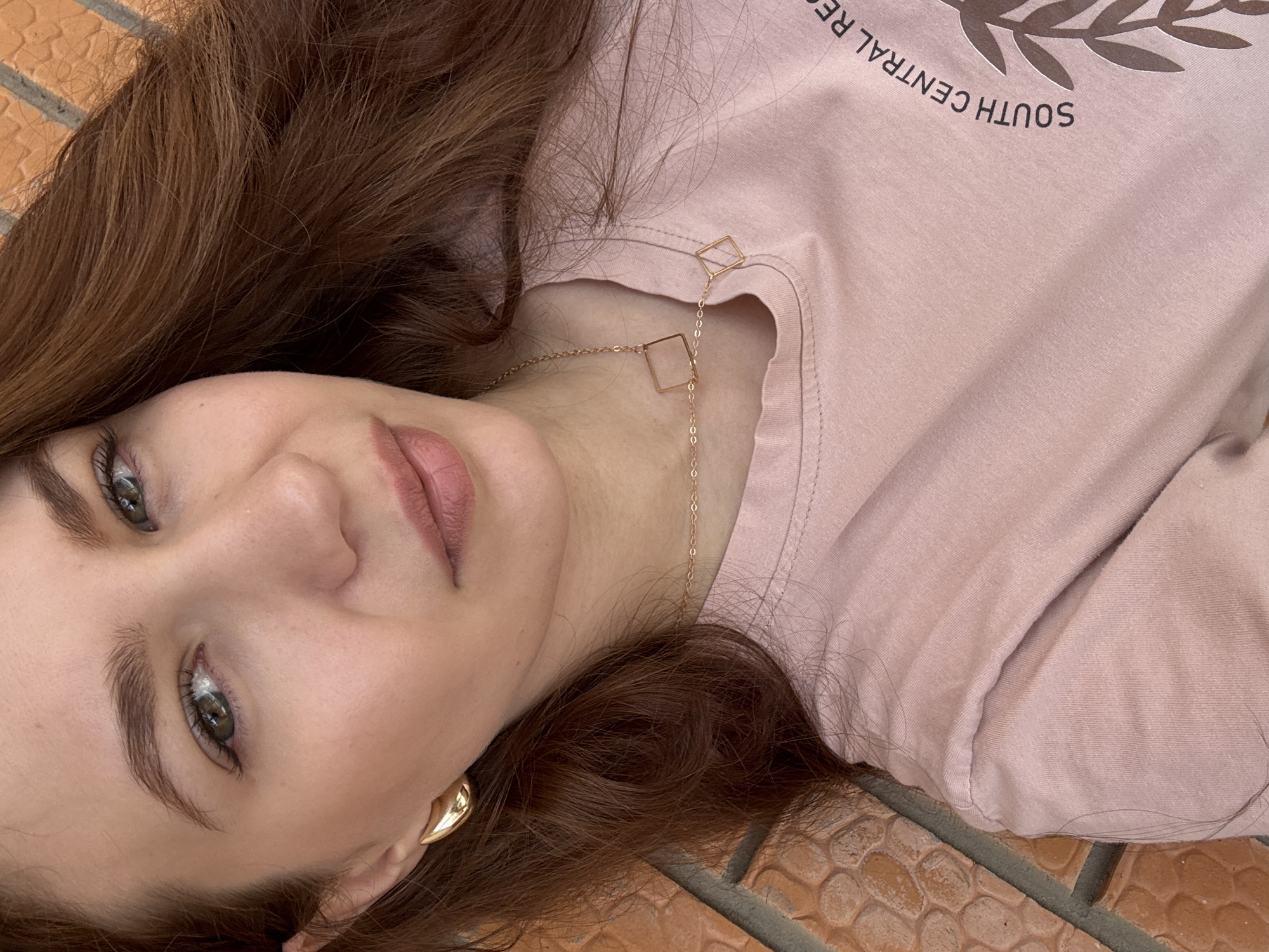 A medium shot photo shows a young woman lying down on a textured surface, possibly a brick wall or a tiled floor. She is looking towards the camera with her head tilted. Her skin is fair, and she has green eyes and dark, wavy hair that is spread around her. She is wearing a light pink t-shirt with a faded graphic print, which appears to include the words "SOUTH CENTRAL RES." and a leaf motif. She is also wearing a gold earring in her visible ear and a delicate gold necklace with two square pendants. The background is a reddish-orange textured surface, possibly bricks. The lighting is natural, illuminating her face and highlighting her features. The location is not specified in the image, and therefore it cannot be determined whether it is in Chaikovsky, Russia.