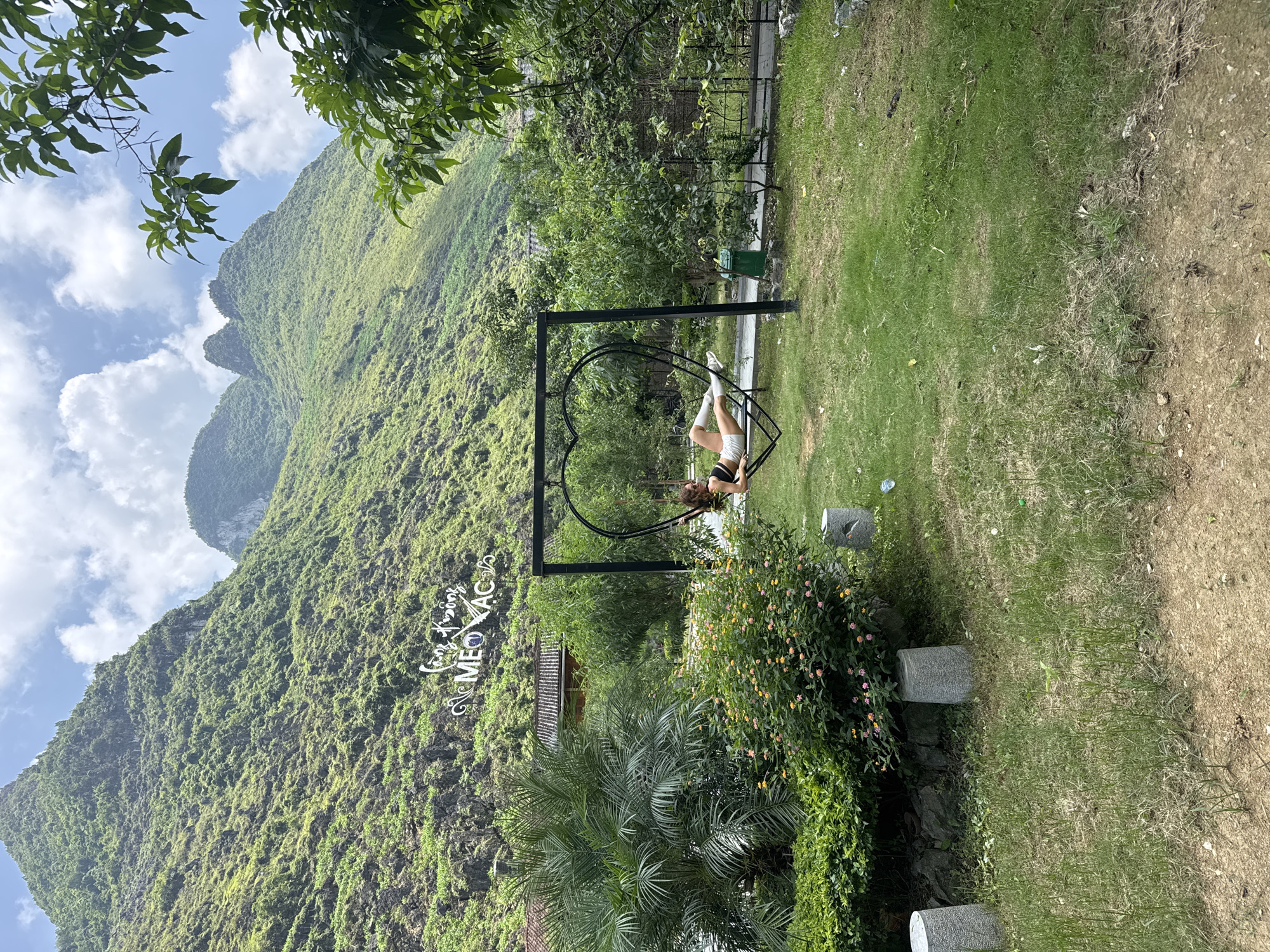 A scenic shot depicts a woman relaxing on a heart-shaped swing in Mèo Vạc Commune, Vietnam. The backdrop features a lush, green mountain, partially obscured by the swing's structure. The woman is reclined, with her legs extended, suggesting a relaxed posture. The surrounding environment is grassy, with various plants and small concrete blocks scattered around, adding a natural feel to the setting. A sign reading "Meo Vac" in Vietnamese is visible on the mountainside.