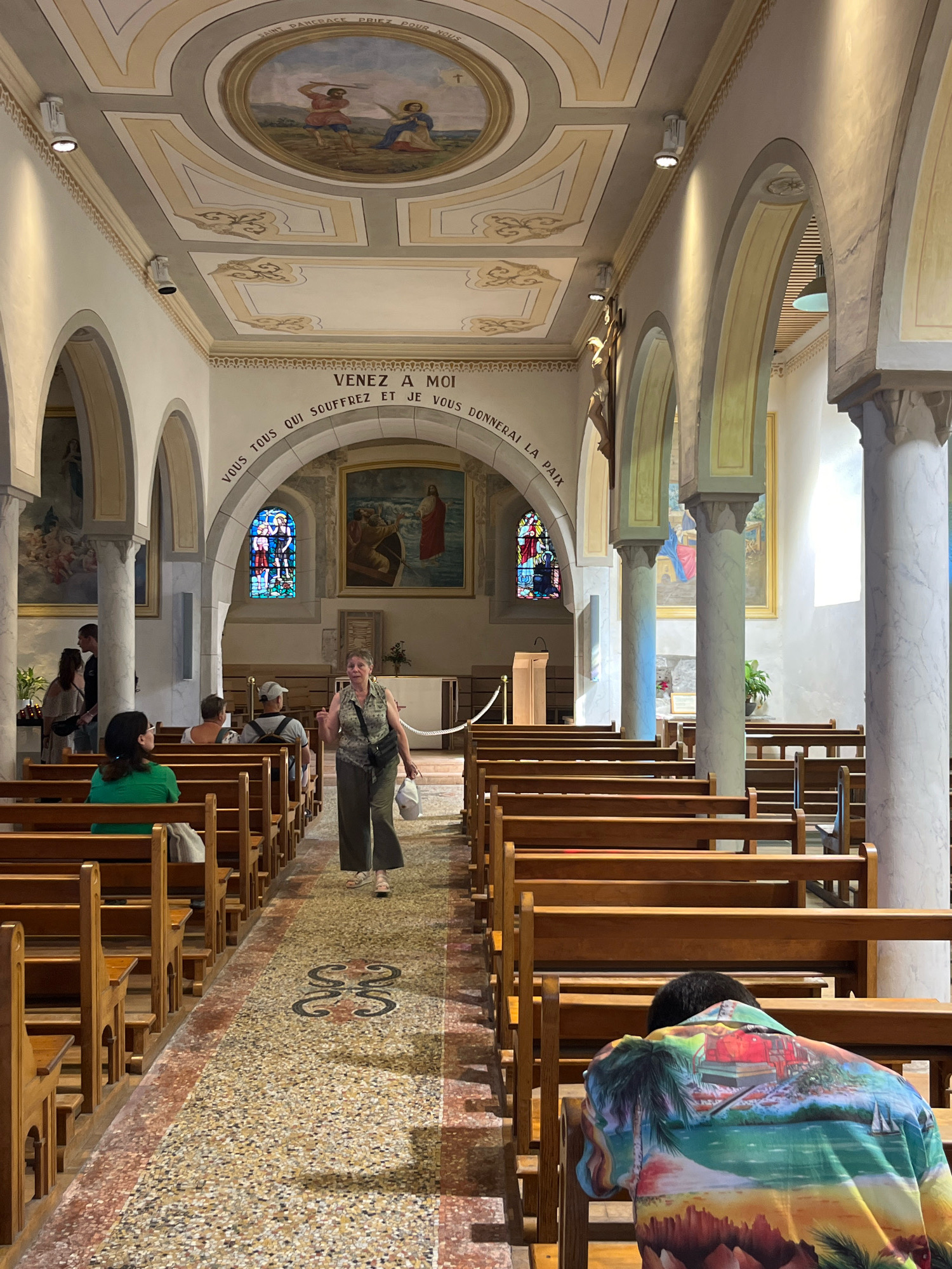 The image depicts the interior of the Saint Pancras Church in Yvoire, France, a Romanesque church dating back to the 12th century. The nave is lined with wooden pews, and the floor is a mosaic of terrazzo tiles. A woman in a floral top and green pants is walking down the aisle, carrying a white bag. In the foreground, a person wearing a colorful Hawaiian shirt is seated on a pew, with their head bowed. The ceiling features a fresco of a biblical scene. Stained-glass windows and paintings adorn the walls. The overall atmosphere is serene and reflective.