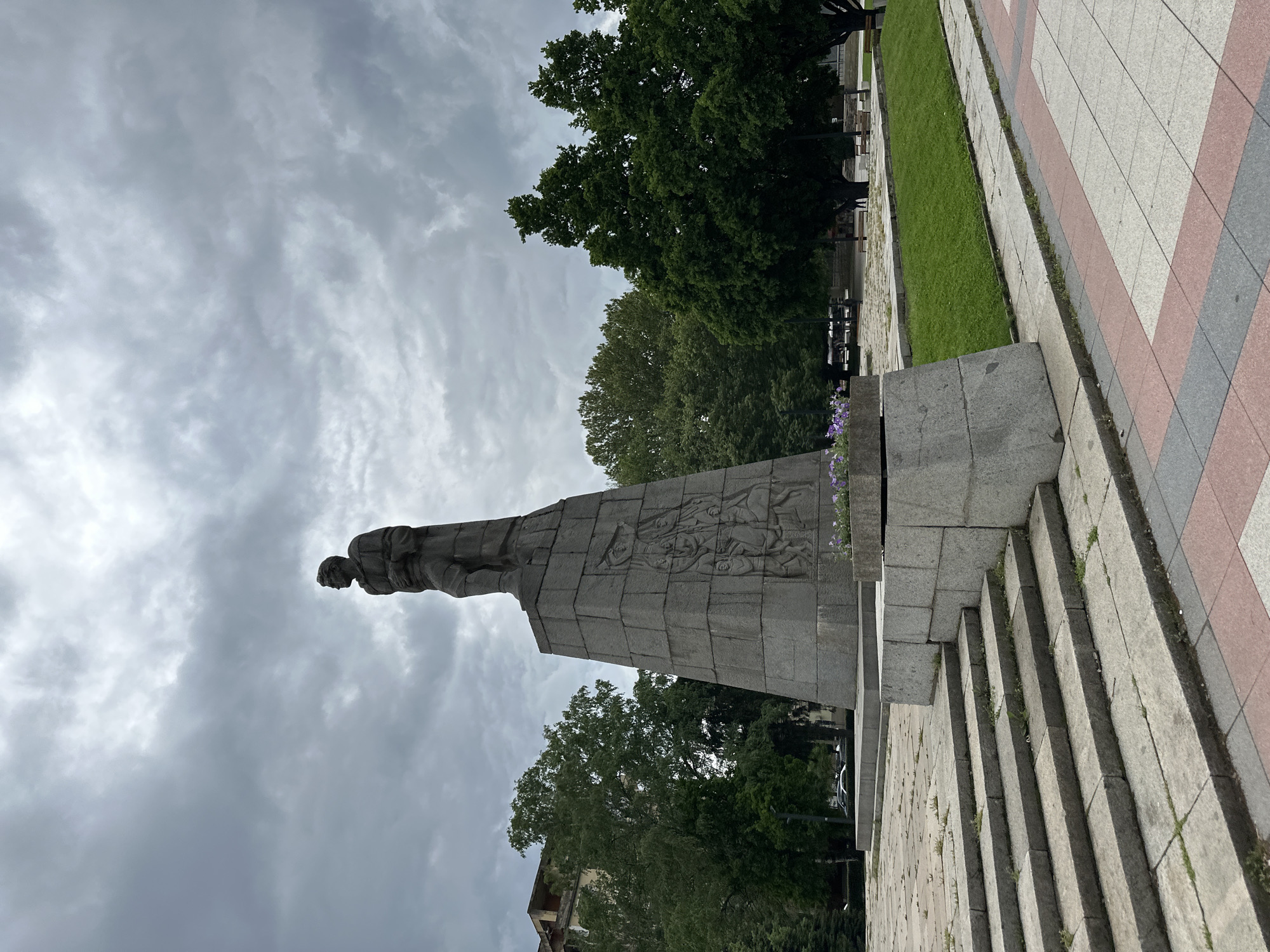 The image shows a monument in Montana, Bulgaria. The monument is made of gray stone and features a male figure atop a tall pedestal, with bas-relief carvings on its sides. The sky is overcast with heavy clouds. The monument is surrounded by trees and a paved walkway. There are steps leading up to the monument. Grass and benches are visible in the background.