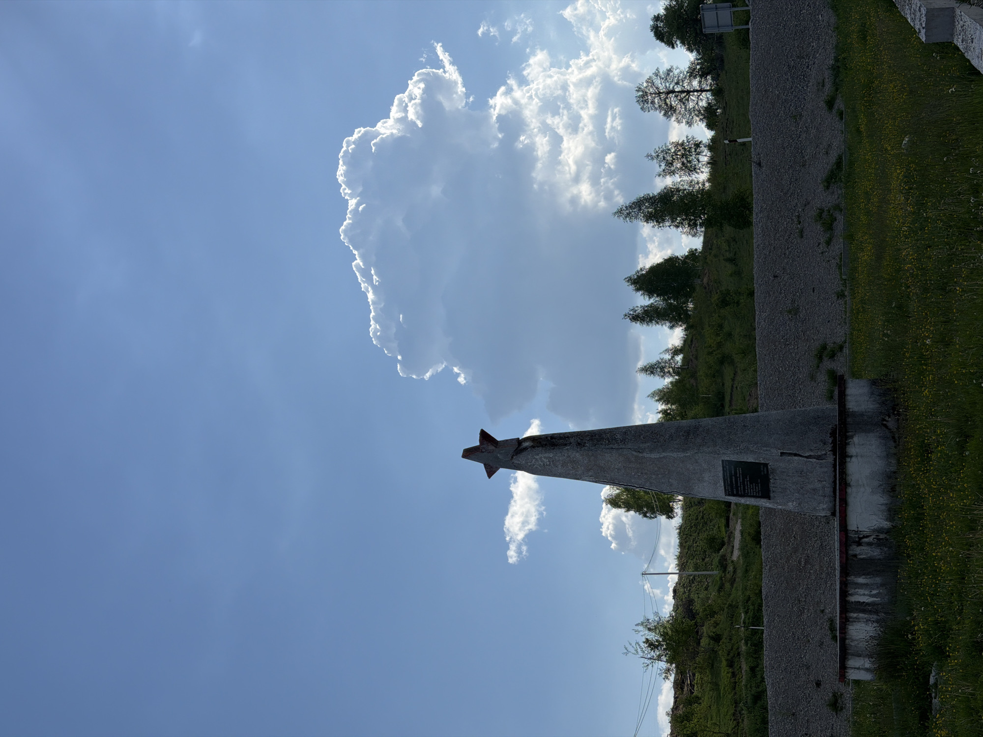 A photograph shows a concrete monument with a five-pointed star on top in Marina Šuma, Montenegro. The monument is triangular, wider at the base, and has a plaque on its side. It is positioned on a concrete base. To the right of the monument is a gravel area with some vegetation. In the background, there are trees and a hill. The sky is blue with a large white cloud on the left side of the frame. The photograph is rotated counter-clockwise.