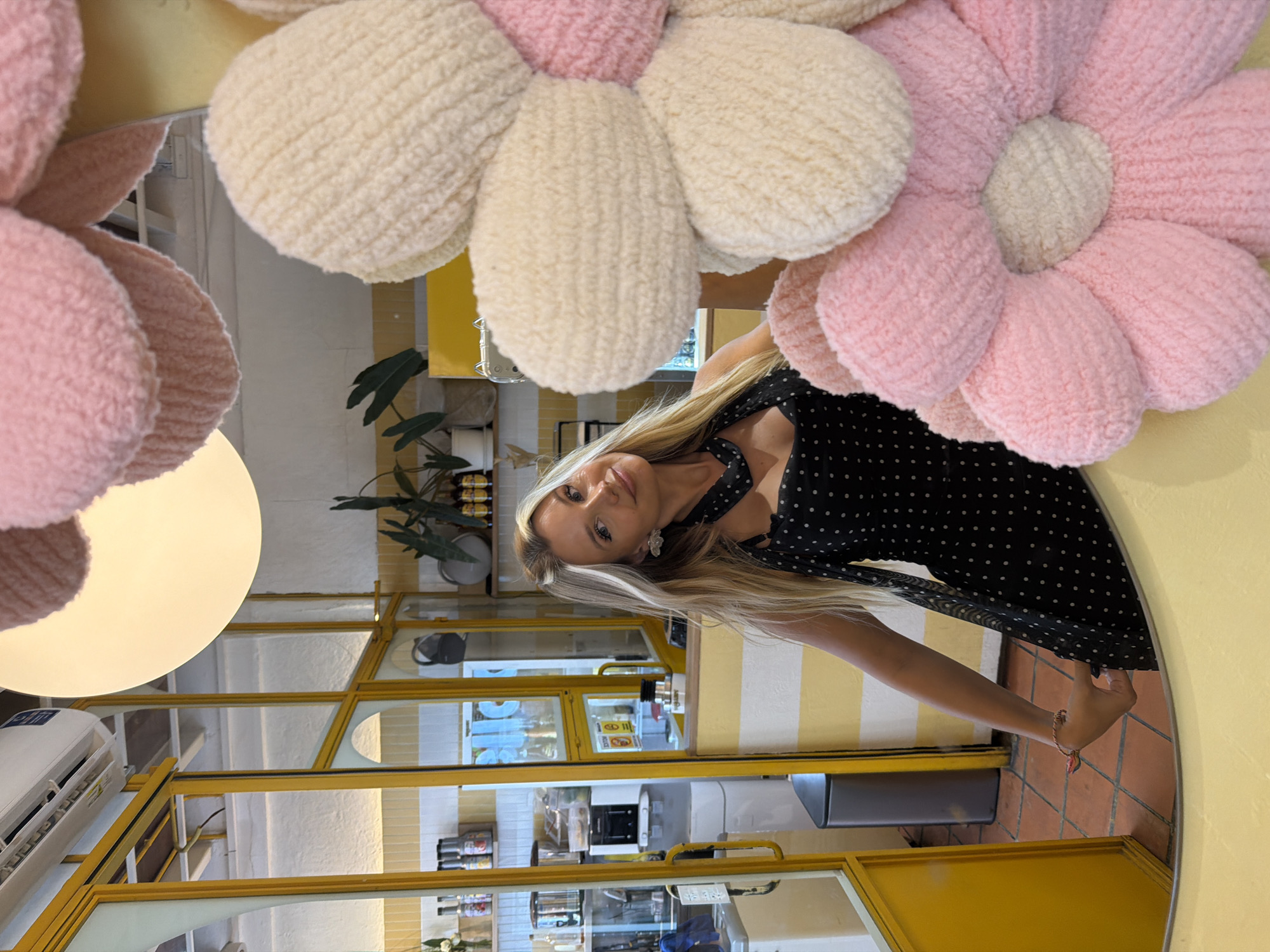 A blonde woman in a black polka dot dress poses in a room with yellow and white striped walls and a bright yellow doorway. She is reflected in a large mirror. The room appears to be a cafe or restaurant, with shelves stocked with bottles and a water dispenser visible in the background. A large, plush flower decoration, made of pink and cream-colored yarn, is in the foreground, partially obscuring the woman and the environment. The woman is wearing a heart-shaped charm bracelet and a white flower earring. The scene is brightly lit, likely by indoor lighting. The location is not specified as being in Phuket City Municipality, Thailand.