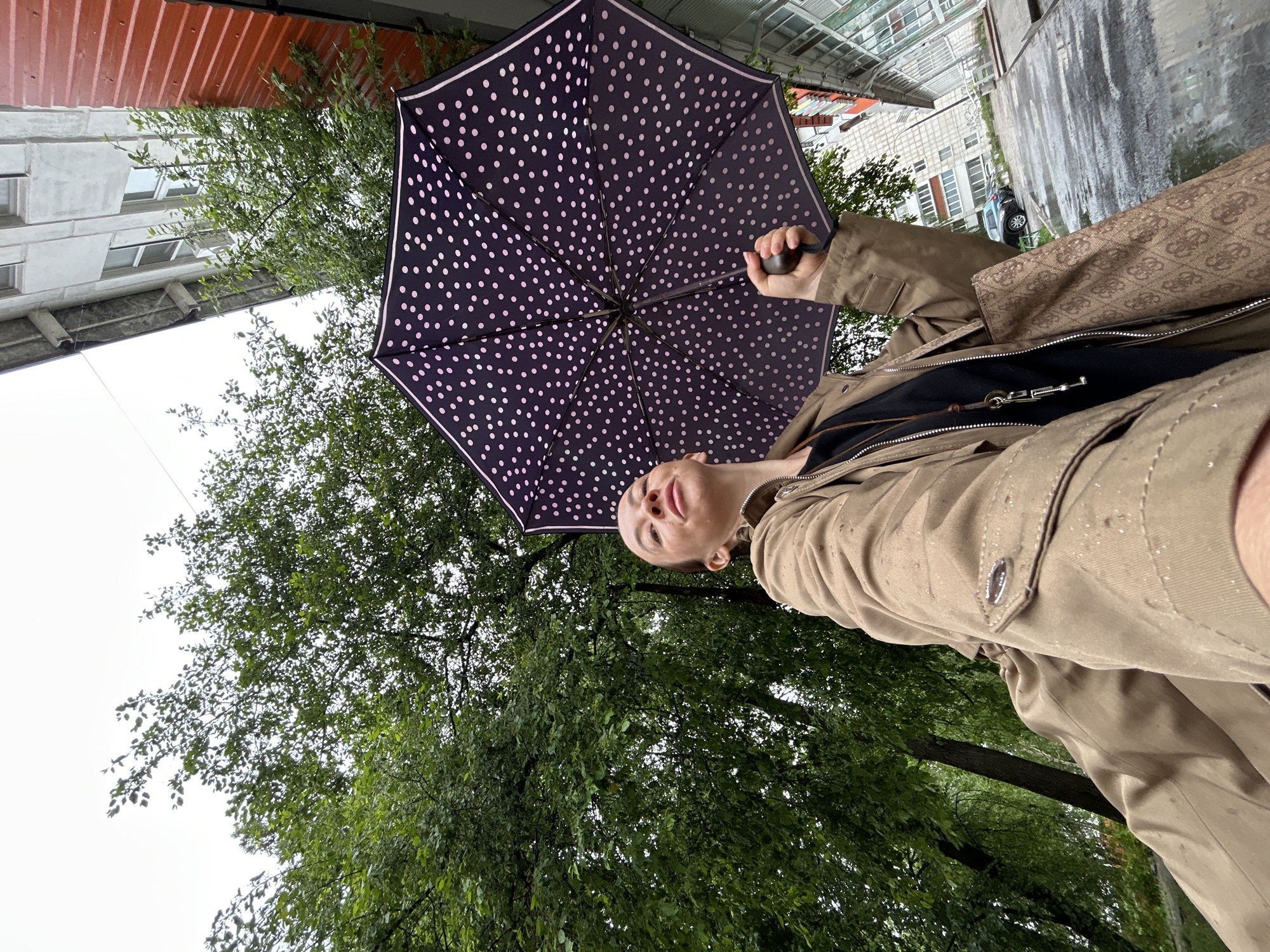 A medium shot depicts a person holding a polka-dot umbrella. The person is wearing a khaki-colored jacket over a black top, and they appear to be looking upwards. The background includes green foliage of a tree. A building is visible in the upper portion of the image. Based on the available information, the location is tentatively identified as the city of Chaikovsky, Russia.