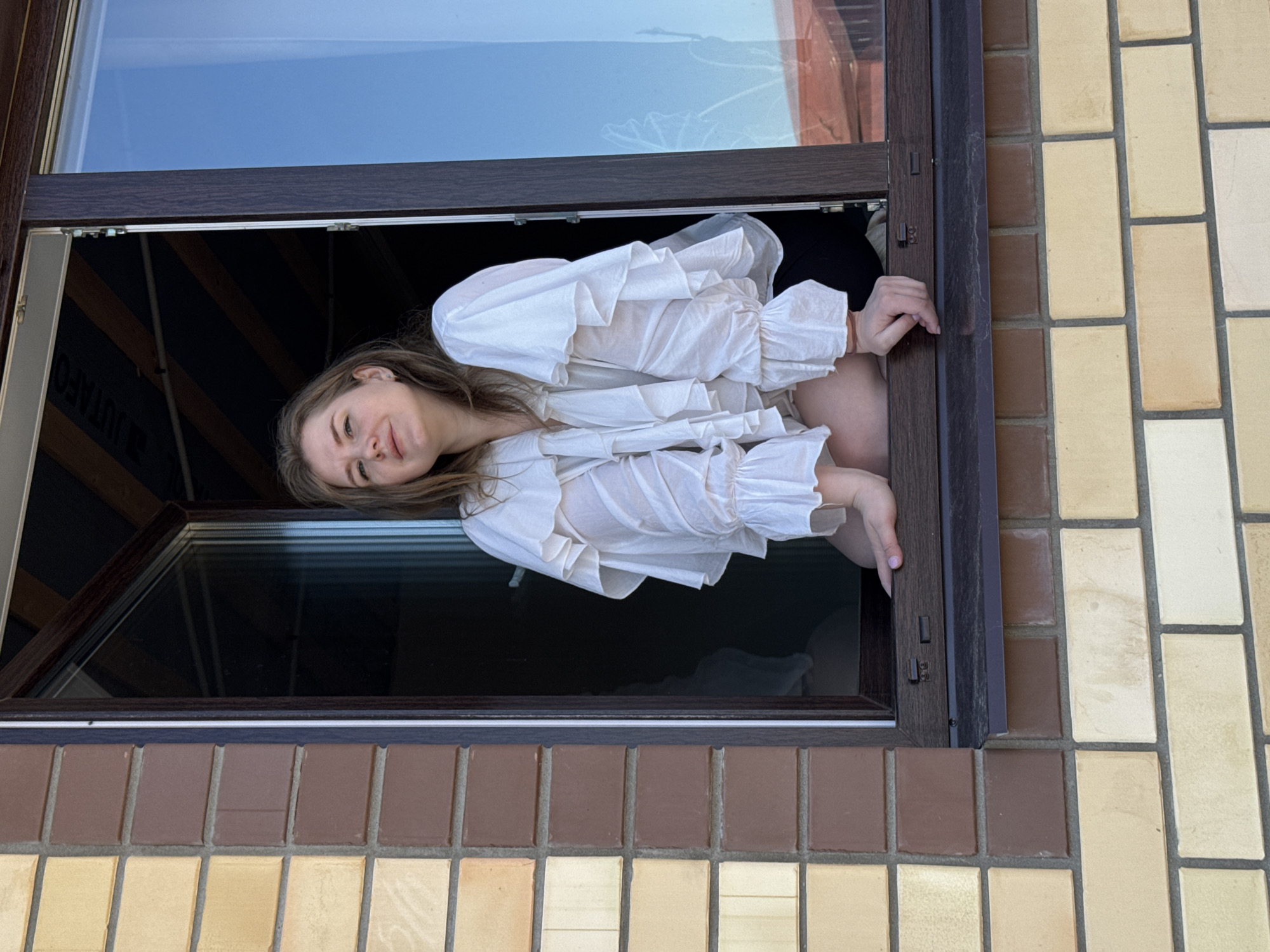 A young woman is leaning out of a partially opened window, wearing a ruffled white shirt. The window is surrounded by brickwork, alternating in color between tan and brown. The location is possibly in Gornyi (Dvurechenskoe S/P), Russia.
