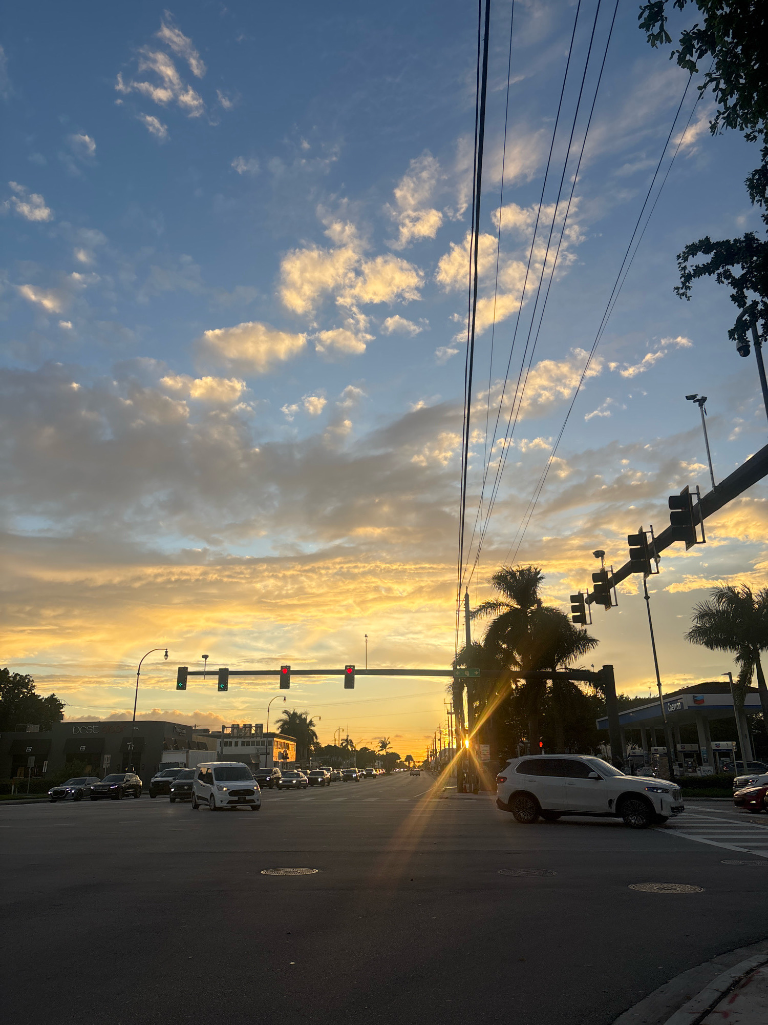 The image depicts a street scene in Hallandale Beach, Florida, United States of America, during a sunset. A traffic light hangs over a multi-lane road where several cars are visible, suggesting moderate traffic. The sky is filled with clouds illuminated by the setting sun, casting a warm, golden glow. Palm trees are present, characteristic of the Florida landscape. Power lines are strung across the upper portion of the frame, and traffic lights are mounted on poles. A Chevron gas station is visible in the background. The overall impression is that of a typical urban environment at dusk, with the natural beauty of the sunset adding a dramatic element to the scene.