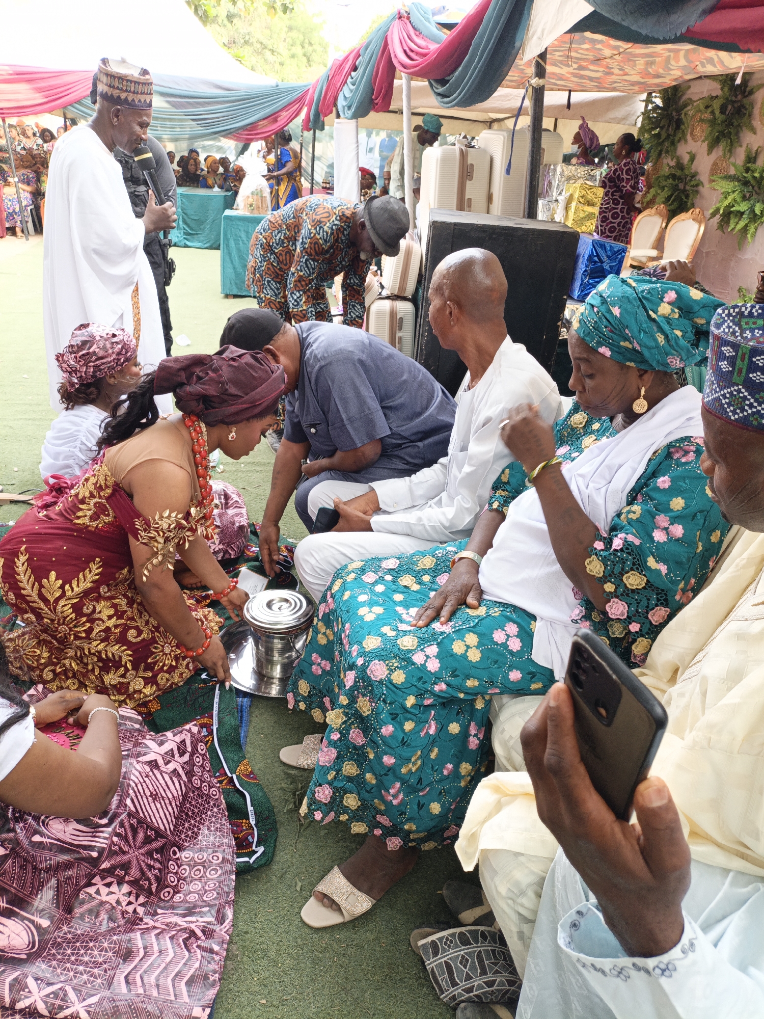 This image captures a vibrant outdoor gathering or ceremony in Sabon Gari West, Nigeria, taking place on a green turf under a colorful canopy draped with pink, blue, and green fabrics. The scene is bustling with people dressed in traditional attire, indicating a significant cultural event.
In the foreground, a woman wearing an elaborate red and gold embroidered dress with a matching head wrap kneels on the ground. She is adorned with prominent red beaded jewelry and appears to be presenting or holding a metal tray containing a silver lidded container. Another person in a dark patterned garment sits beside her.
In the midground, several individuals are seated on the ground. To the right, an older woman with distinctive facial scarification marks wears a teal green lace-patterned dress with pink and yellow floral accents, a white shawl, and gold earrings. She is seated next to a man in a light-colored traditional robe and an embroidered cap, who is holding a smartphone. A man in a white robe and an embroidered cap stands to the left, speaking into a microphone, suggesting he might be an officiant or speaker. Behind the kneeling woman, a man in a blue shirt leans forward, appearing engaged with the central activity.
The background reveals more attendees, some seated, others standing. Large white suitcases and several wrapped gifts in gold, blue, and white are visible, stacked on a platform or table, possibly part of a dowry or presentation. Decorative green foliage also adorns the setting. The bright lighting suggests it is daytime.
Visible text includes "MADE IN NIGERIA 100% COTTON" printed on a white strip along the edge of a green patterned fabric near the kneeling woman. The overall atmosphere appears celebratory and communal, centered around a traditional ritual or presentation.