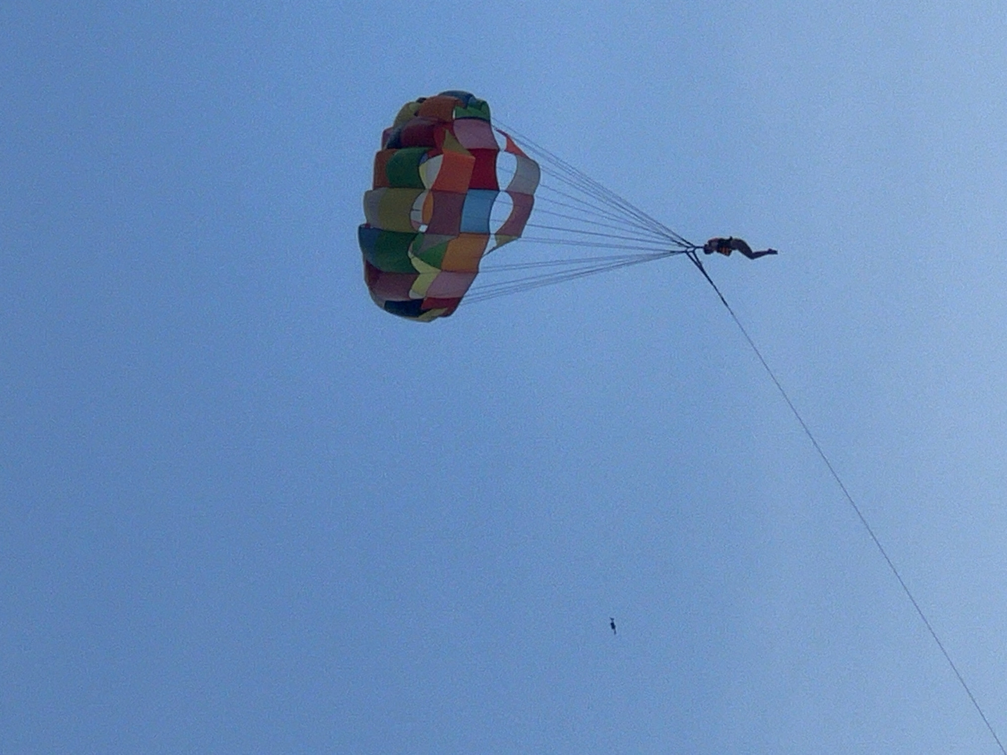 The image shows a person parasailing against a clear blue sky. The parachute is colorful, with panels of red, yellow, green, and blue. A single person is attached to the parachute by cords and is suspended in the air, with their legs dangling. In the distance, a small black object, possibly another parasail or a small aircraft, is visible in the sky. The scene suggests an outdoor recreational activity, likely taking place near a coastal area, which is common in Da Nang, Vietnam, known for its beaches and water sports.