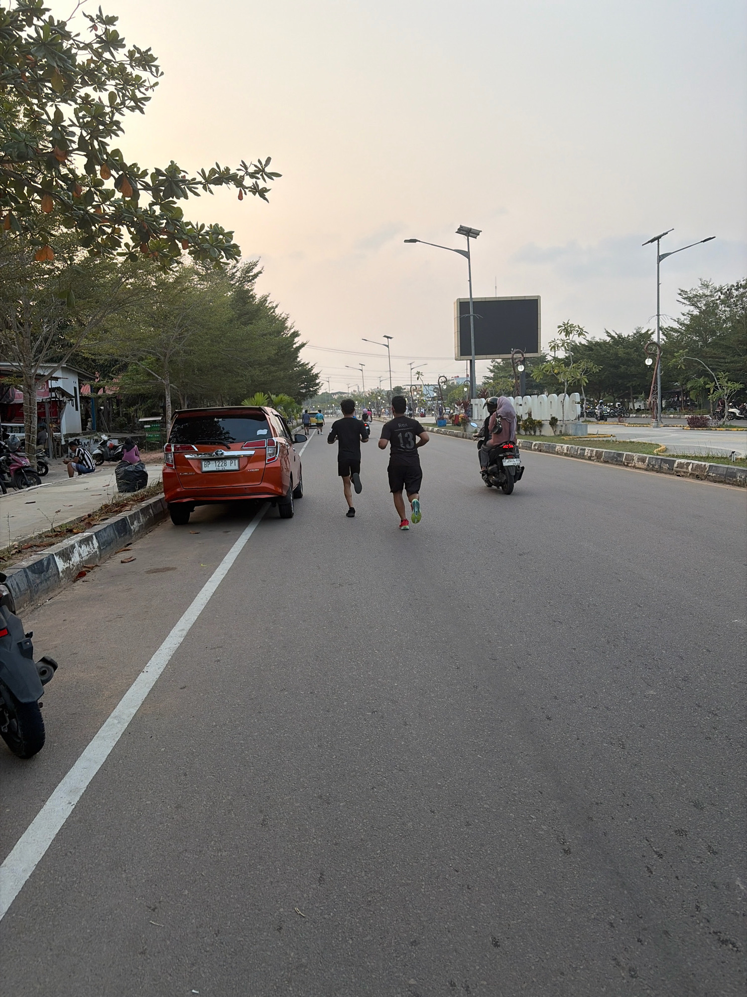 Two men are jogging on a paved road in Tanjung Pinang, Indonesia, on a street lined with trees. One man, wearing a black t-shirt with the number "13" on the back and black shorts, is in the foreground, running with his arms pumping. The other man, in a black t-shirt and shorts, is slightly ahead and to the left. A red car is parked on the side of the road to the left of the joggers. Further down the road, a motorcycle with two people on it is passing by. In the background, there are streetlights, a large digital billboard, and more trees. The sky is overcast with a pale yellow hue. On the left side of the road, there are a few small shops and parked motorcycles.