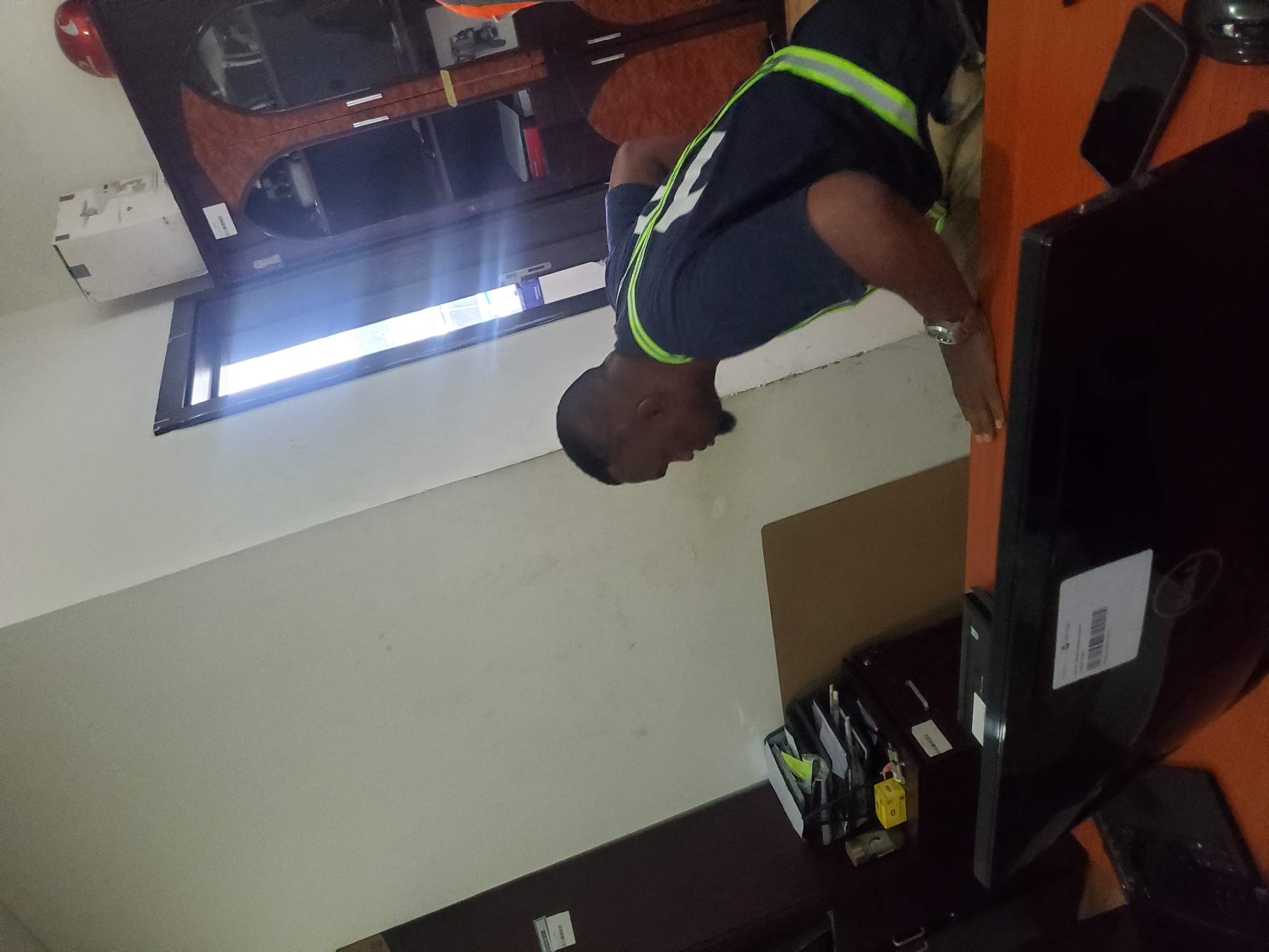 An indoor scene, likely an office or workspace in Gwagwada, Nigeria, captured during the daytime with bright ambient light. The primary subject is a dark-skinned man with short hair, wearing a dark short-sleeved shirt adorned with bright green/yellow reflective stripes. He is seen leaning forward with his head down, suggesting focus or engagement with something on the desk in front of him. A silver-colored watch is visible on his left wrist.
The setting includes a wooden desk with a reddish-brown top and dark base. On the desk to the right, a black flat-screen computer monitor stands, next to which lies a black smartphone. A desk organizer containing various stationery items and yellow tags, one appearing to show the number "0," is also on a darker table surface in the foreground. A white rectangular box or unit, possibly an air conditioning unit, is mounted on a light-colored wall. A strong light source emanates from a window or opening on the left side of the frame, framed by dark sills. Another piece of furniture, possibly a wooden cabinet or shelving unit, is visible in the background, topped by a red object that might be a helmet or part of equipment.
Visible text includes the word "INVENTORY" along with some numbers (partially obscured, possibly "0033") on a white label attached to a structure near the air conditioning unit. A barcode and some illegible text are also present on a label on the back of the monitor. The bright lighting suggests it is daytime.