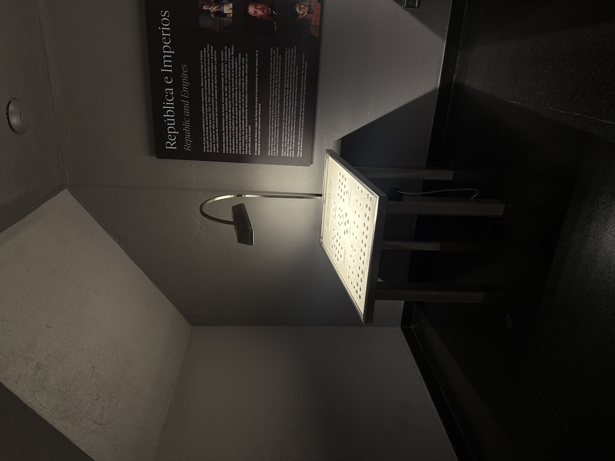 The image depicts an exhibit in a dimly lit room, possibly a museum in San Pedro Garza García, Mexico, based on the Spanish language signage. On the left wall, a dark gray information panel titled "República e Imperios / Republic and Empires" features a description in Spanish and English about the history of copper coins in Mexico. Below the panel is a white table with small, dark, round objects displayed on its surface, illuminated by a gooseneck lamp. The floor is dark gray, with a slight sheen, possibly polished stone. The surrounding walls are light gray and converge into a corner. No people are visible.
