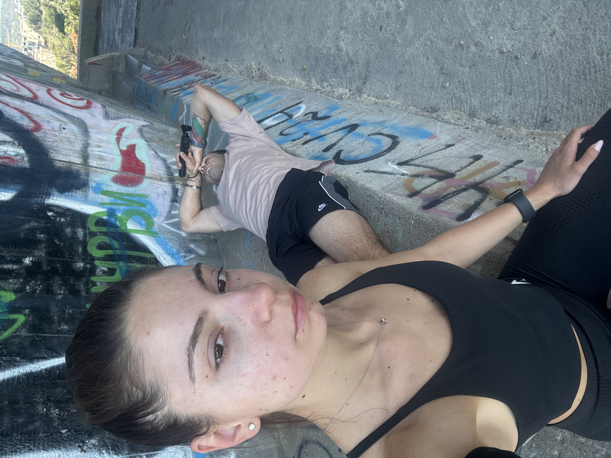 The image depicts a selfie taken by a young woman in the foreground, with a man lying down in the background. The woman is wearing a black sports bra and black leggings, and has her hair pulled back. She has a necklace with a small heart pendant and earrings. The man is wearing a light-colored t-shirt and black athletic shorts, and appears to be resting his head on his arms, with his eyes closed. He is holding a black cylindrical object in his right hand.
They are situated outdoors, on a concrete surface next to a wall covered in graffiti. The environment suggests an urban setting, possibly a park, overlook, or an underpass. The graffiti on the wall includes various colors and tags. In the background, a city skyline with buildings and greenery is visible, which, combined with the style of architecture and vegetation, suggests the location is likely Varna, Bulgaria, possibly from an elevated viewpoint. The overall atmosphere is casual and relaxed.