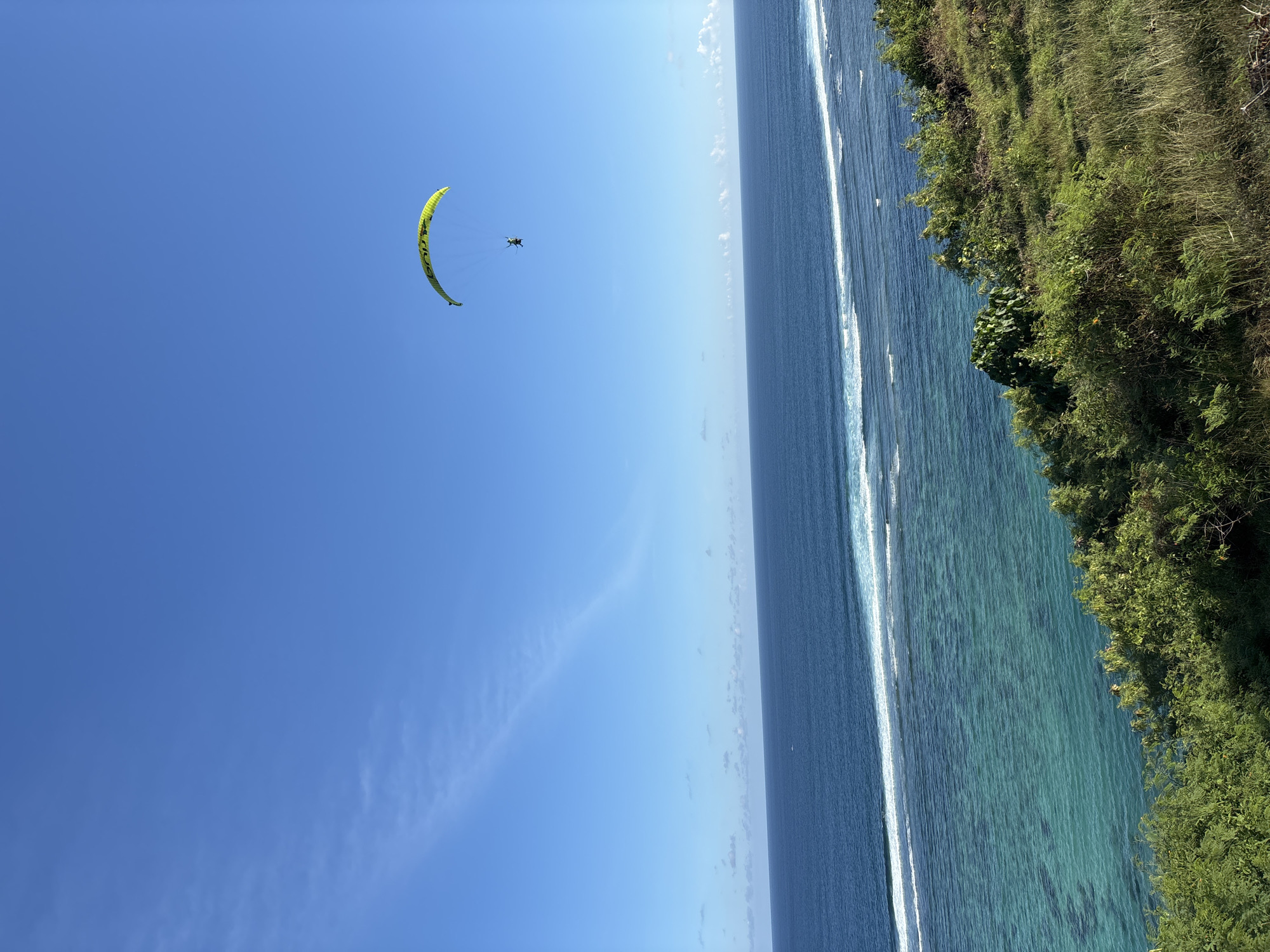 A person is paragliding in a blue sky over the sea in Unknown, Indonesia. The sea has white waves, and there is green vegetation on the coastline.