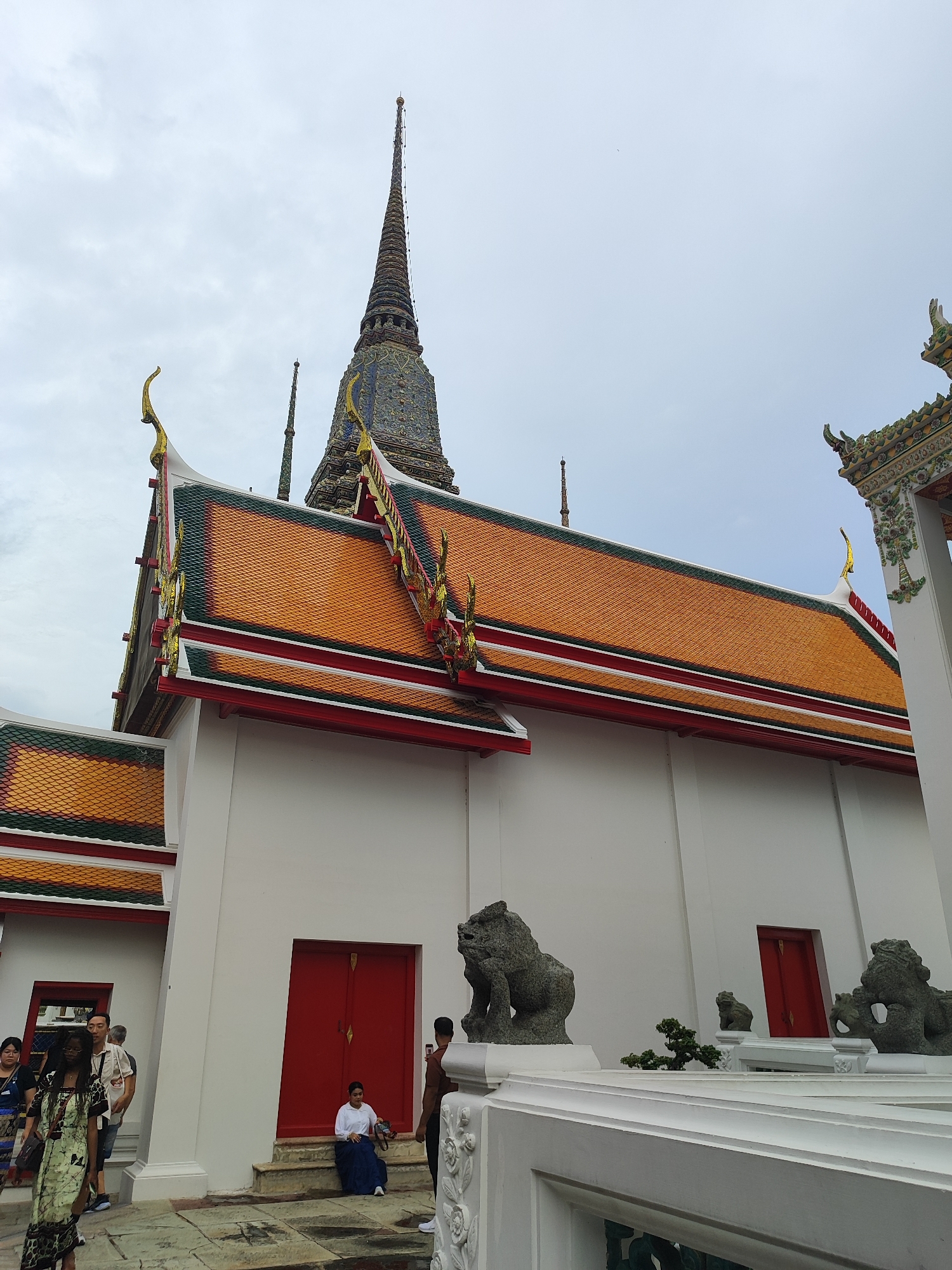The image shows a side view of a white building with an orange tiled roof, adorned with ornate gold trim. A tall, multi-tiered spire, characteristic of Thai Buddhist temples, rises in the background, partially obscured by the building. Several people are present, including a woman in a patterned dress walking in the foreground, and another woman sitting on the steps of the building. Stone lion statues, a common architectural element in Southeast Asian temples, are visible on a white railing in the foreground. The scene appears to be in a temple complex in Bangkok, Thailand, given the architectural style.