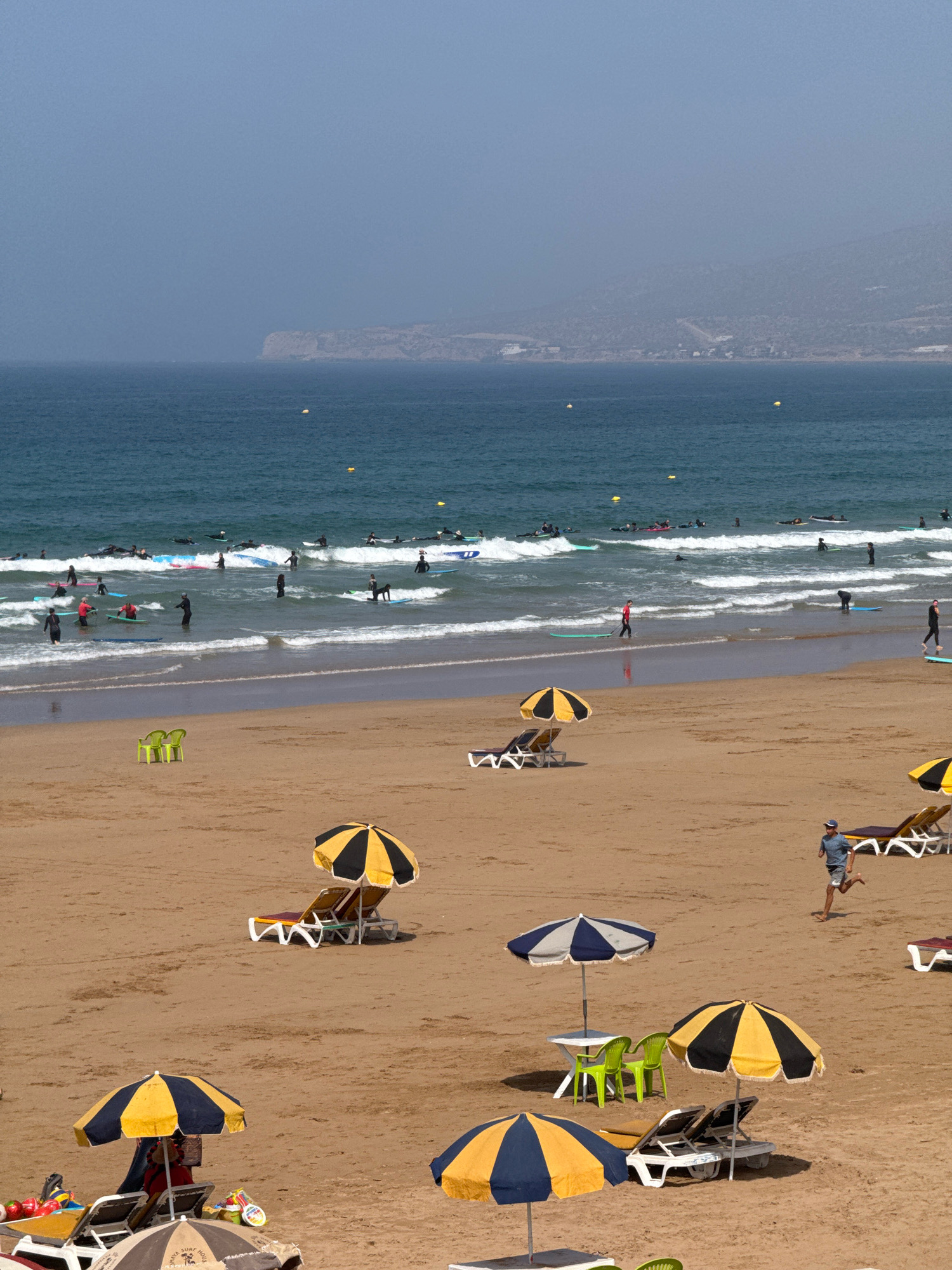 The image depicts a wide, sandy beach in Aourir, Morocco, under a hazy sky. The ocean in the background is a deep blue, with numerous surfers and their boards visible in the waves. Several yellow buoys are scattered in the water. The shoreline is populated with people, some wading, others on surfboards. The beach itself features rows of empty lounge chairs and umbrellas, some open and providing shade, others folded. A few green plastic chairs and a small white table are set up on the sand. In the foreground, a man in a blue shirt and shorts is running across the beach. There are also various beach accessories like bags and umbrellas visible. The overall scene suggests a busy day at the beach with recreational activities taking place in both the water and on the sand.