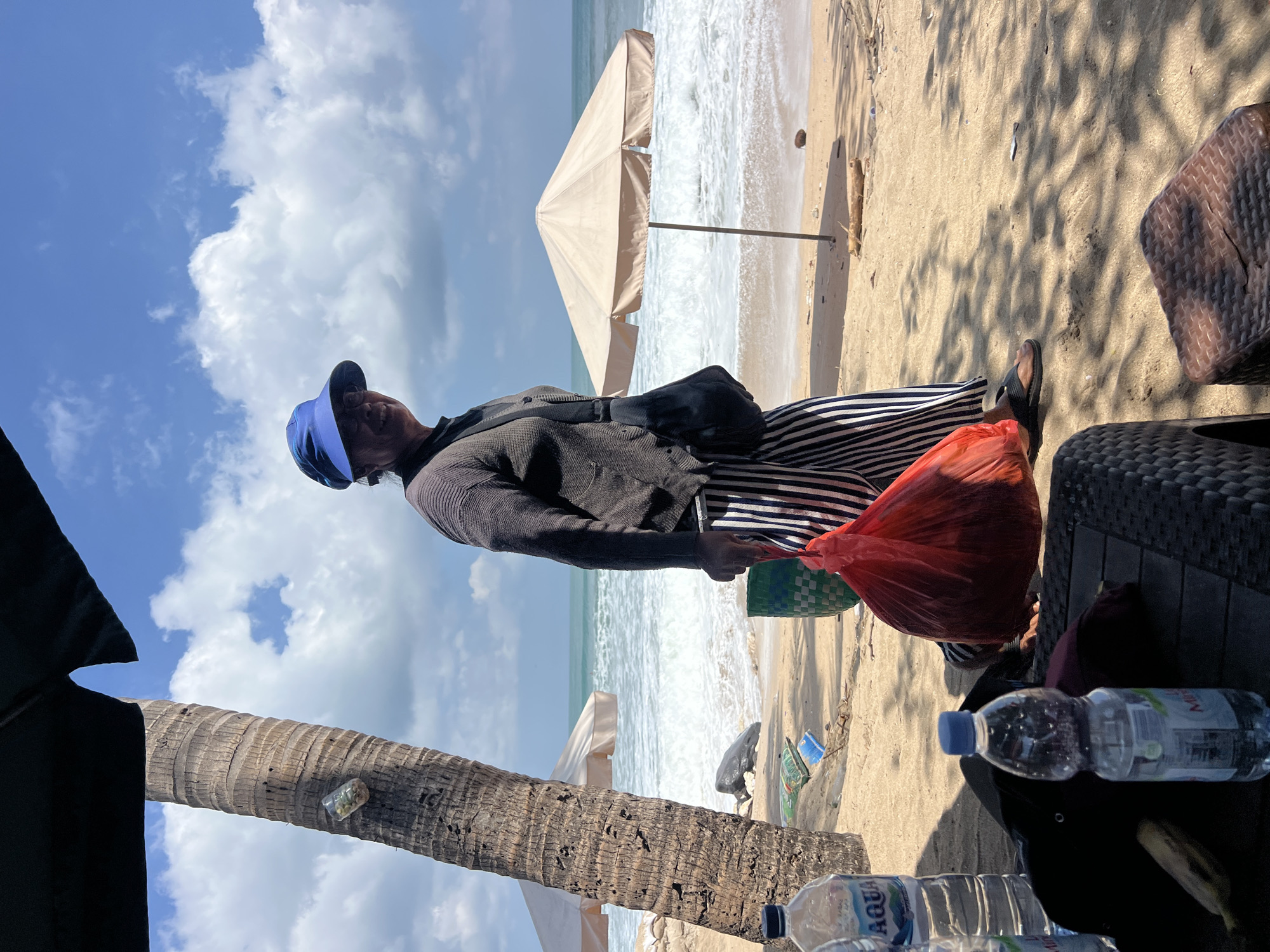A woman wearing a blue sun hat and a dark long-sleeved shirt with vertical black and white striped pants walks on a sandy beach in Kuta, Indonesia. She is carrying a large, red plastic bag in her right hand and has a dark bag slung over her left shoulder. The woman is smiling and looking towards the viewer. Behind her, the ocean has white-capped waves under a bright blue sky with scattered clouds. A tan beach umbrella stands near the water's edge. In the foreground, a palm tree is visible, with a clear plastic cup containing small objects attached to its trunk. Various bottles of water and a wicker table are also in the foreground, suggesting a seating area on the beach. Debris, including a green basket and some discarded items, is scattered on the sand. The lighting indicates it is daytime, with shadows cast by the palm tree and overhead canopy.