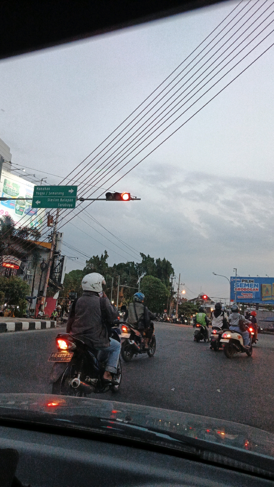 The image captures a street scene in Surakarta, Indonesia, characterized by a traffic light showing red, indicating a halt. Several motorcycles are stopped at the intersection, with their riders wearing helmets. In the foreground, the dashboard of a car is visible, suggesting the perspective of a vehicle occupant. Overhead, a dense network of power lines stretches across the sky, partially obscured by a gray, overcast sky. Billboards and street signs are present on the sides of the road, with one green sign indicating directions to "Manahan," "Yogya / Semarang," and "Stasiun Balapan Surabaya." The overall atmosphere suggests a typical urban commute during twilight hours.