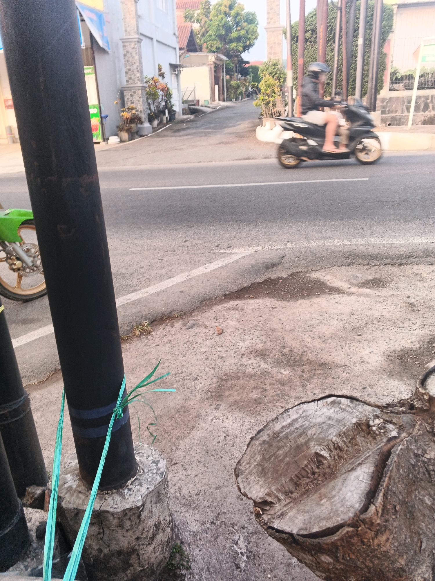 The image captures a street scene in Semarang, Indonesia. In the foreground, a large black pole stands vertically, partially obscuring a bright green motorcycle parked on the left. The pole is secured to a concrete base with turquoise plastic ties. Further back, a person wearing a black jacket and helmet rides a black motorcycle with gold wheels, moving from right to left across the asphalt road. The road is marked with a white line. On the right side of the road, a tree stump with visible rings lies on the ground. The background features buildings, trees, and a fence, indicating an urban environment. The lighting suggests it is either early morning or late afternoon.