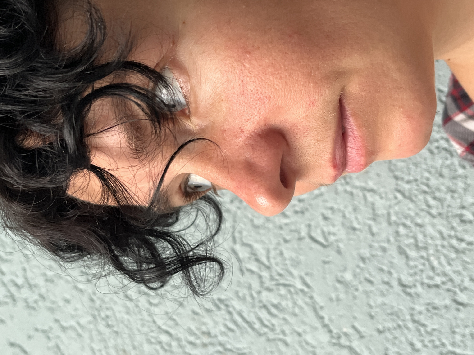 The image shows a close-up, profile view of a person's face against a textured, light blue background. The person has dark, curly hair that frames the face, and is looking to the left. Their skin is visible with details of pores and some redness around the nose and cheeks. The lighting suggests an outdoor setting, possibly during the day, casting a soft light on the face. The background appears to be a wall with a rough, stucco-like texture. No specific city or location details are discernible from the image itself, and therefore, no association with ღრმაღელის თემი, Georgia can be made.
