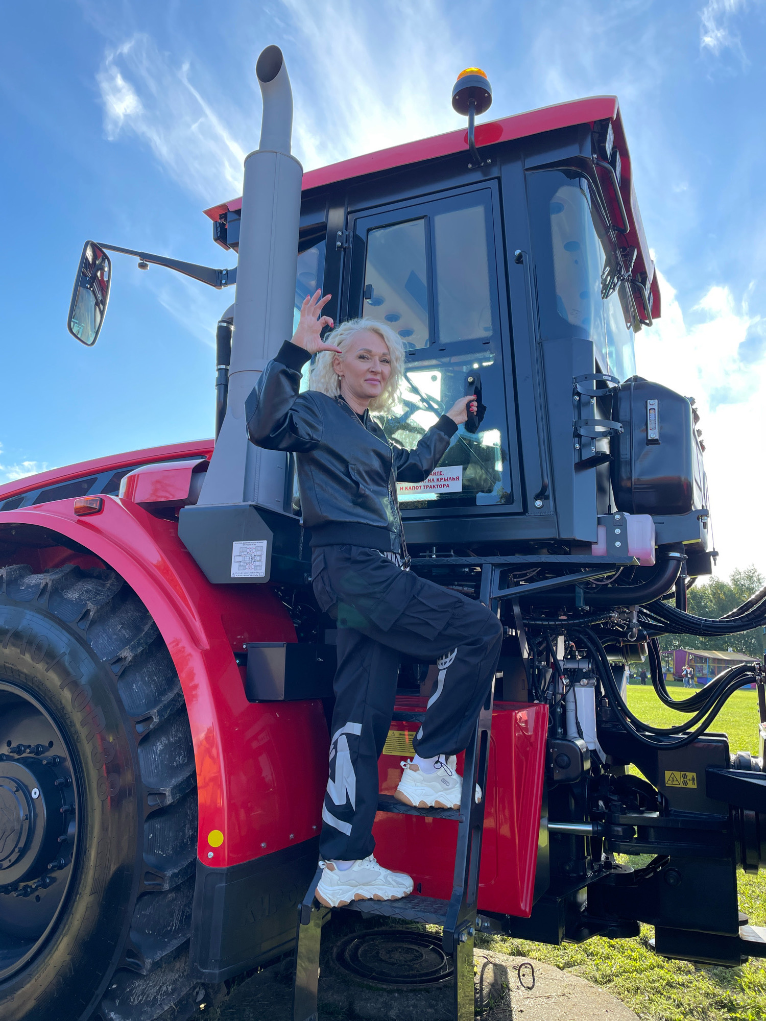 A blonde woman wearing a black leather jacket and black cargo pants with white accents stands on the steps of a large red tractor. She is facing the camera and making a "peace" sign with her right hand. The tractor has a large exhaust pipe on the side of the cab, and a yellow flashing light on top of the cab. The scene appears to be outdoors on a sunny day with a blue sky and some white clouds. In the background, there is green grass and some distant figures of people and amusement park attractions, suggesting a public event or exhibition, potentially in a park or open field within Saint Petersburg, Russia. The overall impression is one of posing for a photograph at an agricultural or industrial event.
