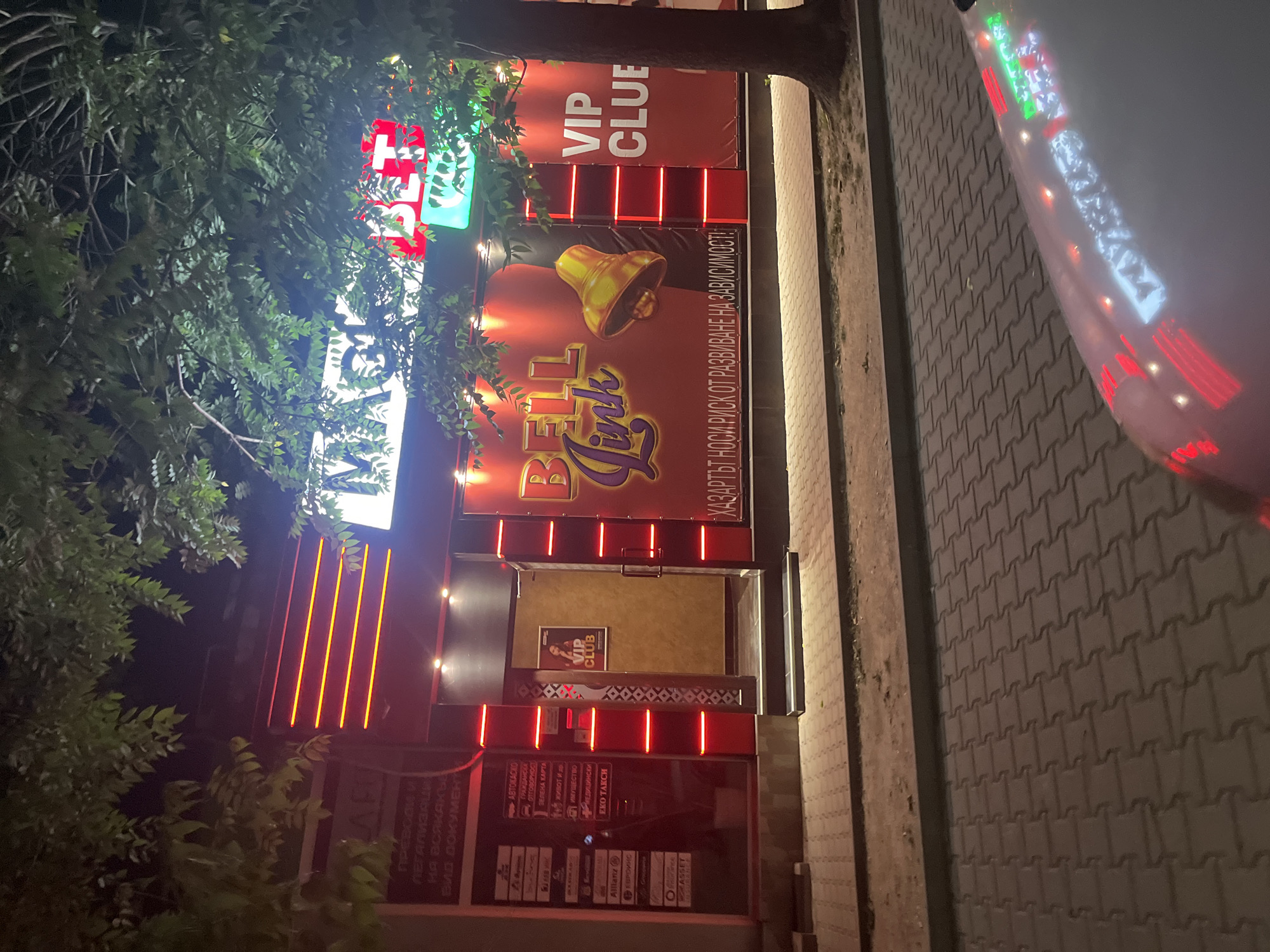 The image depicts the exterior of a building with a prominent sign advertising "BELL Link VIP CLUE" in Blagoevgrad, Bulgaria. The sign features a large bell graphic and text in Bulgarian warning about the risk of developing addiction. The building's facade is decorated with red neon lights. Below the "BELL Link" sign, there are several advertisements, including insurance companies like Allianz and Evroins, as well as services such as "ECO TAXI." There is a VIP Club sign on the front door. In the background, there's foliage, suggesting the building is situated near trees or greenery. Part of a car is visible in the right side of the image.