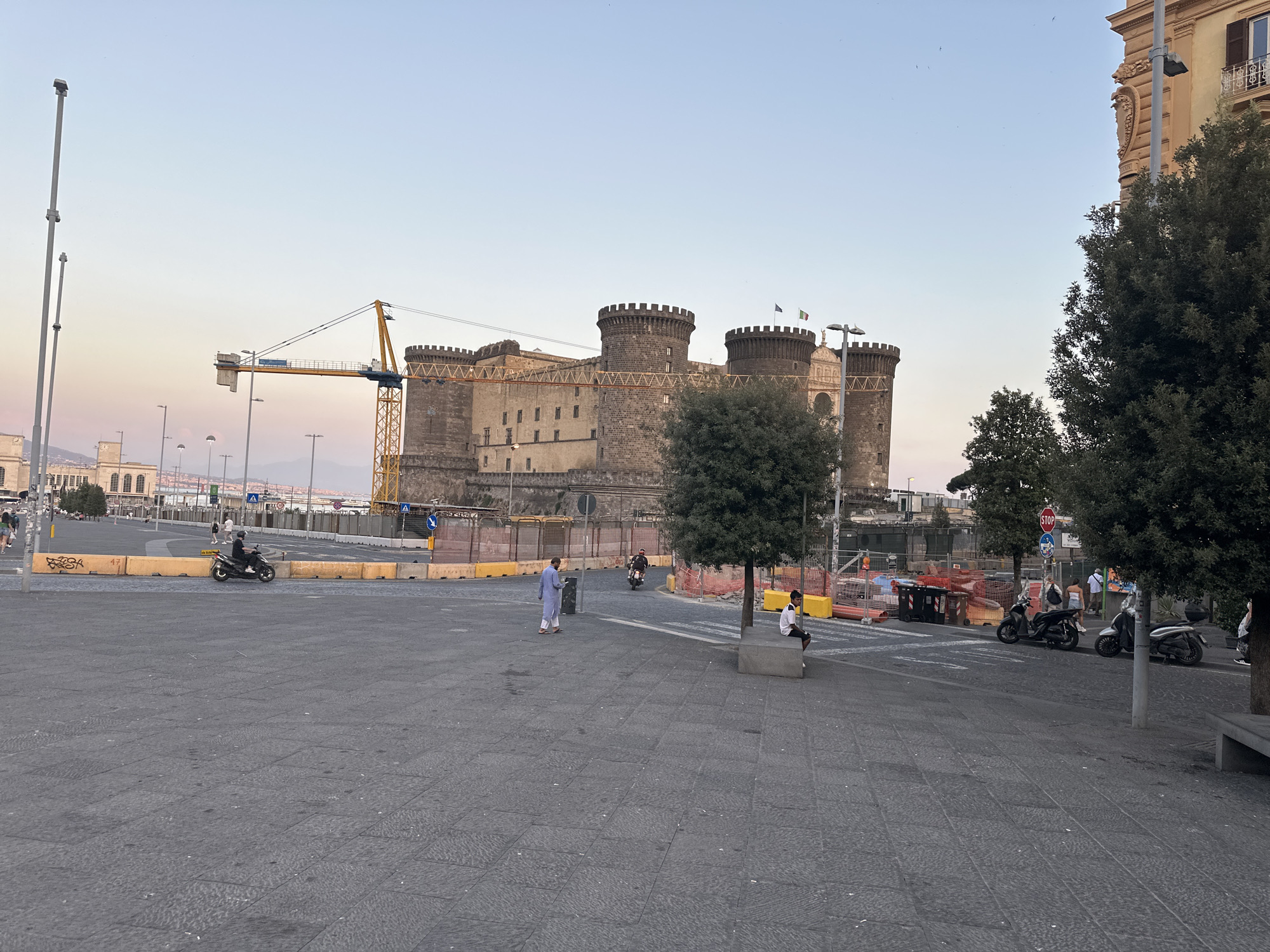 This image depicts Castel Nuovo, also known as Maschio Angioino, in Naples, Italy. The castle is a medieval fortress with multiple towers and a light-colored stone facade. A yellow construction crane is visible to the left of the castle, suggesting ongoing work or development in the area. The foreground is a paved public square with cobblestone patterns. Several people are present in the square. On the left, a person wearing a light blue robe walks across the square. A scooter with a rider is moving from left to right in front of yellow safety barriers. Further to the right, two scooters are parked, and a few people are standing near them. A stop sign is visible on a pole. Trees are scattered throughout the scene, with a large tree in the mid-right foreground and other trees lining the background. The sky is a clear, pale blue, with the soft hues of dusk or dawn visible on the horizon. The overall impression is a city square in front of a historic landmark, with elements of modern urban life and ongoing construction.