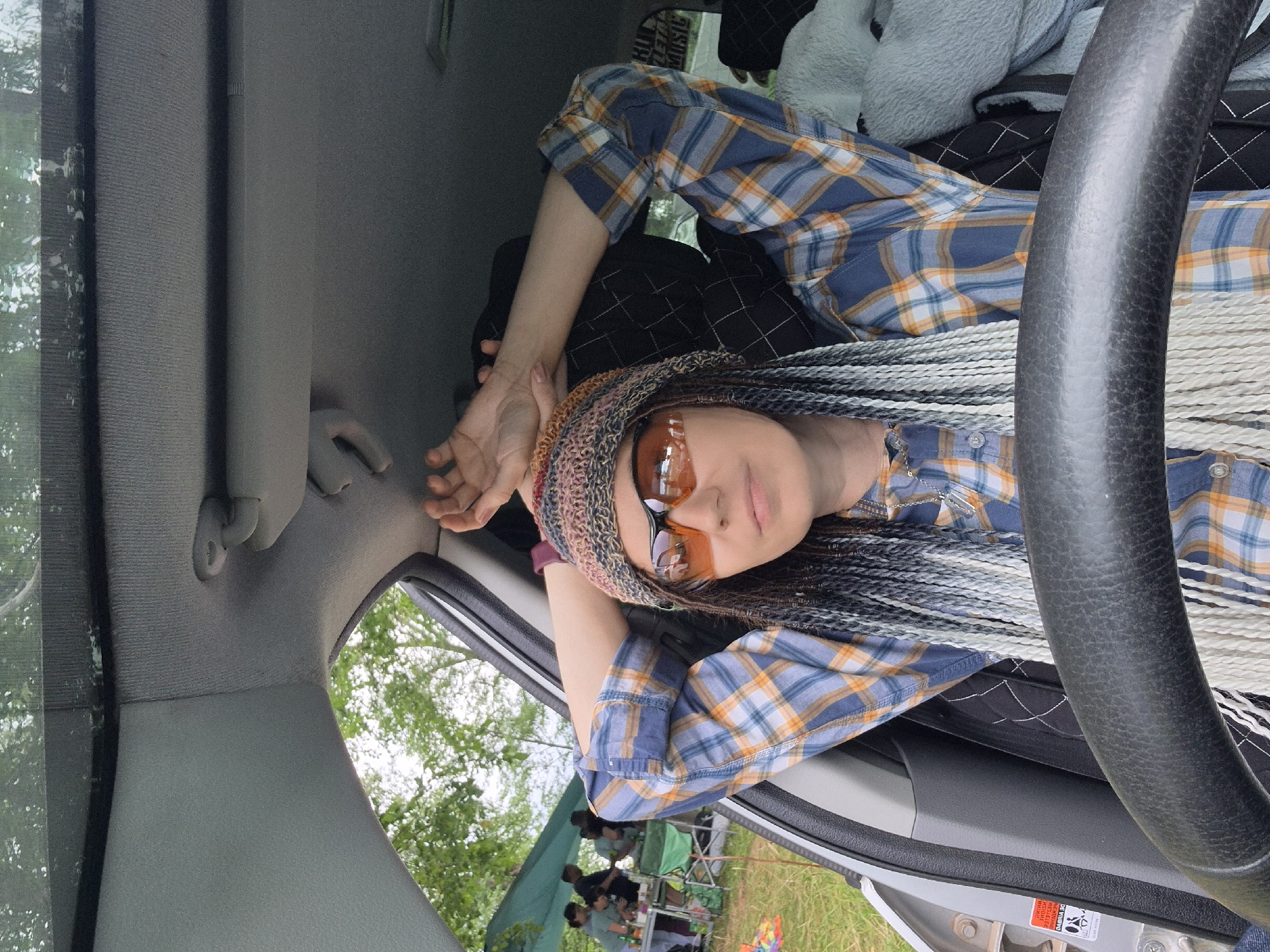 A medium shot captures a woman from the chest up, seated inside a car, her arms resting above her head. She is wearing a blue and orange plaid shirt, a colorful knitted hat, and orange-tinted sunglasses. Her hair is styled in numerous long, thin braids in varying shades of white and black. The interior of the car is visible, including a portion of the steering wheel and the car's roof lining. Through the open car door and windows, an outdoor scene is visible with a green tent, people gathered around tables, and trees in the background. This scene appears to be set in Noviy, Russia, likely at an outdoor gathering or event.