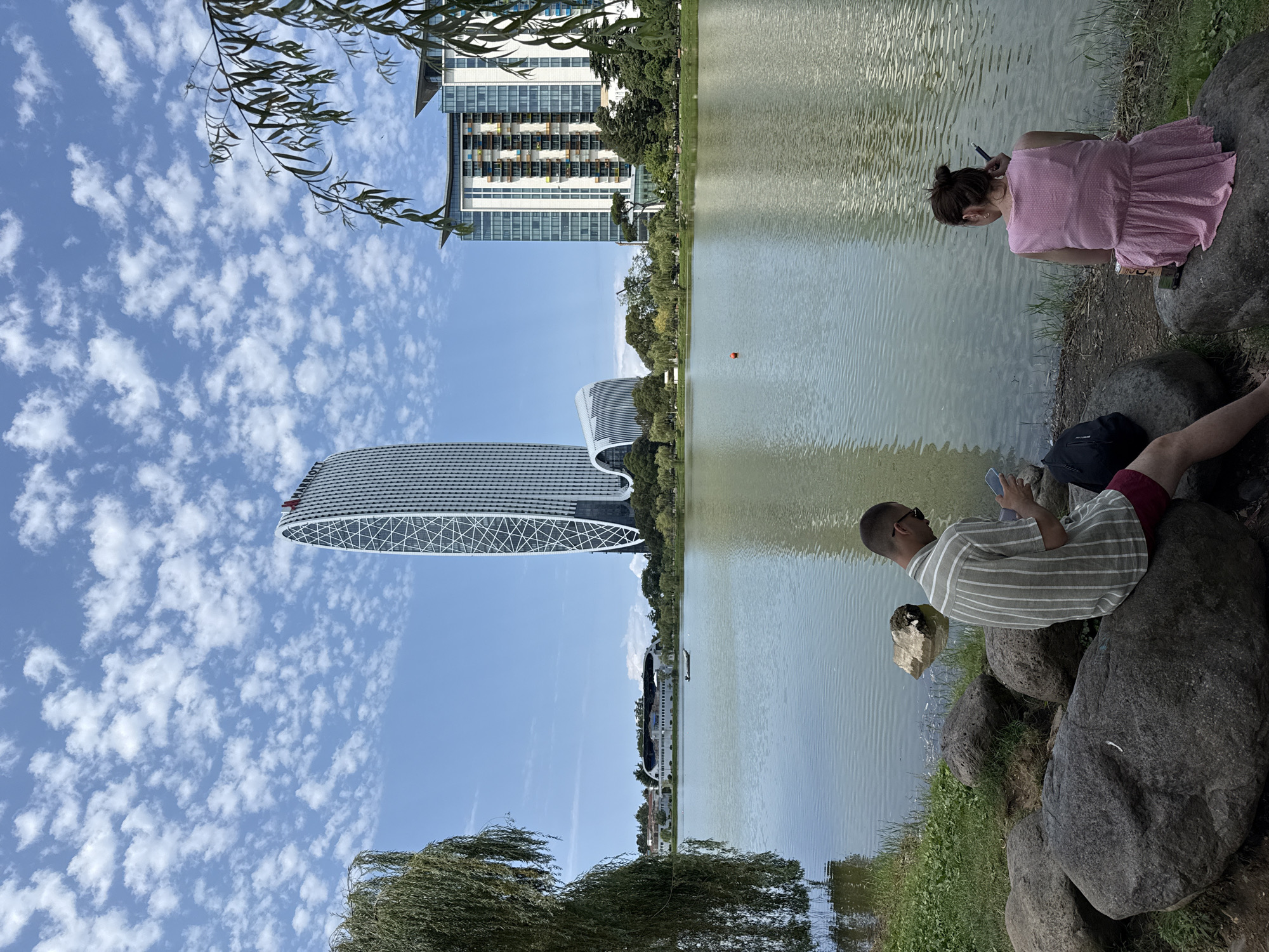 This is a full, eye-level, outdoor, daytime shot of a lake with two individuals seated on rocks in the foreground, facing away from the camera. The man on the left, wearing a vertically striped shirt and red shorts, is looking at his phone. The woman on the right, in a pink dress, is holding a cigarette and a pen. A black backpack is on the ground next to the man, and a beverage can is beside the woman. In the middle ground, the lake extends to a treeline, with a small boat visible in the distance. Beyond the trees are several modern buildings, including a tall, curved skyscraper with a distinctive white lattice design and a lower, rectangular building with a colorful facade. The sky is blue with scattered white clouds.