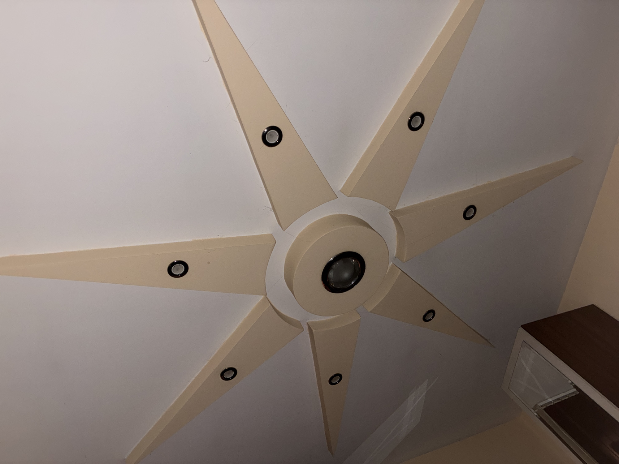This image captures a close-up, low-angle view of a modern ceiling design in an indoor setting, likely in Varna, Bulgaria. The central focus is an elaborate, star-shaped ceiling fixture made of light beige or cream-colored material, set against a white ceiling. The fixture features a prominent circular center, from which eight tapered "rays" extend outwards, resembling a sunburst or an eight-pointed star.
Each of the eight rays, as well as the central circular element, is fitted with a recessed spotlight. These spotlights have a black outer rim and a reflective inner surface. A total of nine lights are visible: one larger light in the center and one smaller light embedded in each of the eight radiating points. The overall design suggests a contemporary and decorative lighting fixture.
In the bottom right corner of the frame, a portion of a piece of dark brown wooden furniture with a glass front is visible, possibly a cabinet or display unit, contrasting with the light ceiling. The artificial lighting from the fixtures and the absence of natural light sources suggest it might be evening or a room with controlled lighting. There are no people or specific activities depicted. Subtle dust or small fibers are visible on the white ceiling surface in some areas. No text is visible in the image.
