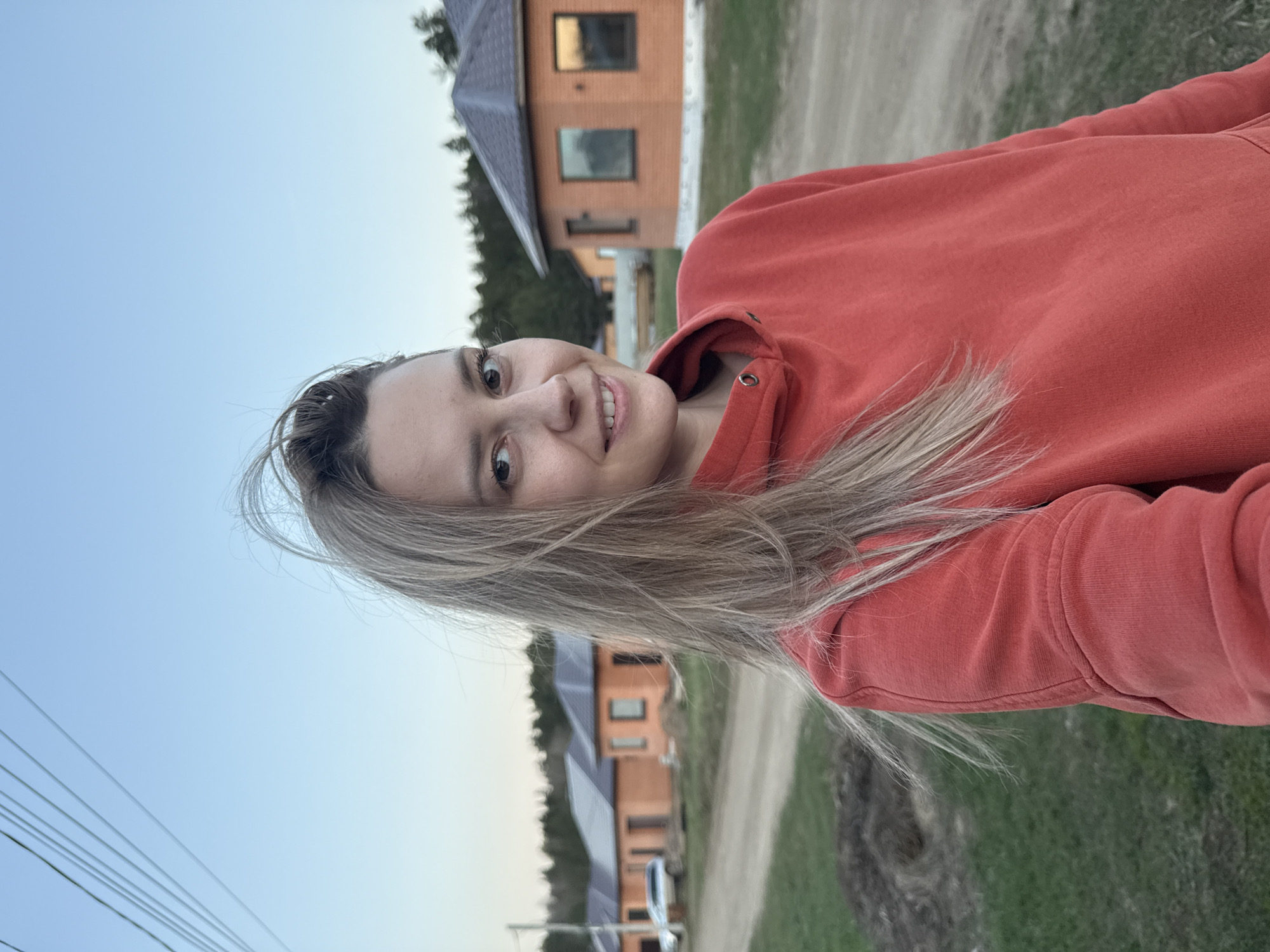 A woman wearing an orange hoodie is standing outdoors. Behind her are newly constructed brick houses with dark roofs and visible windows. A dirt road leads past the houses. The sky is light blue. Power lines are visible in the lower-left corner. This image appears to be taken in a residential area with new construction, but without additional context, I cannot definitively place the location in Chaikovsky, Russia. I would need more data points to make a positive ID.