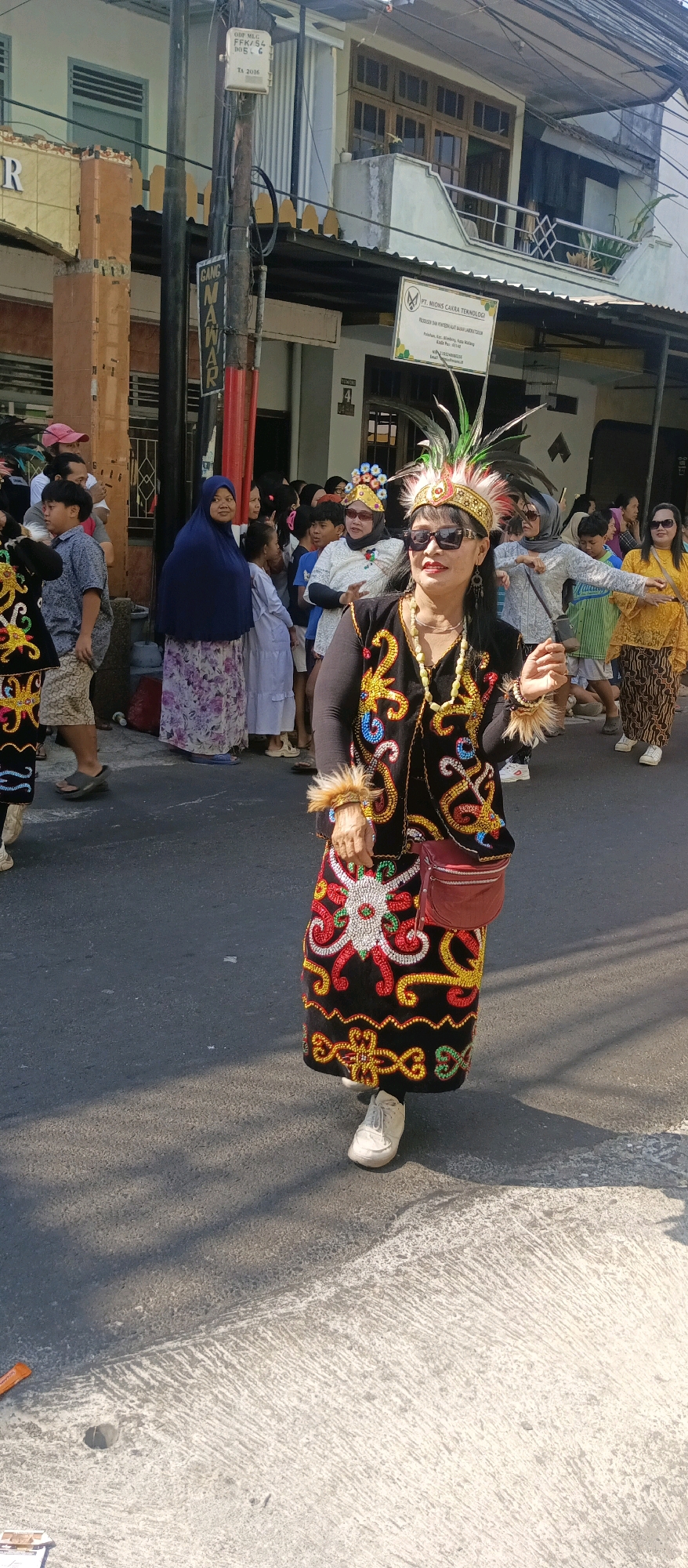 An outdoor daytime photograph captures a woman in traditional attire walking on a paved street. She wears a black garment adorned with vibrant, colorful embroidery in red, yellow, green, and white, forming intricate patterns on the vest and skirt. A red purse is slung across her body. She has dark hair, wears sunglasses, and a feathered headdress with gold accents. Other people, including children and adults, are visible in the background, some appearing to be part of a procession or celebration. Buildings line the street, with one bearing a sign with text that includes "PT. MIONIS CAKRA TEKNOLOGI." The scene suggests a cultural event or festival taking place in an urban setting, likely in Malang, Indonesia, given the presence of the Indonesian building name and the style of the attire. The lighting indicates it is a sunny day, casting shadows on the street.
